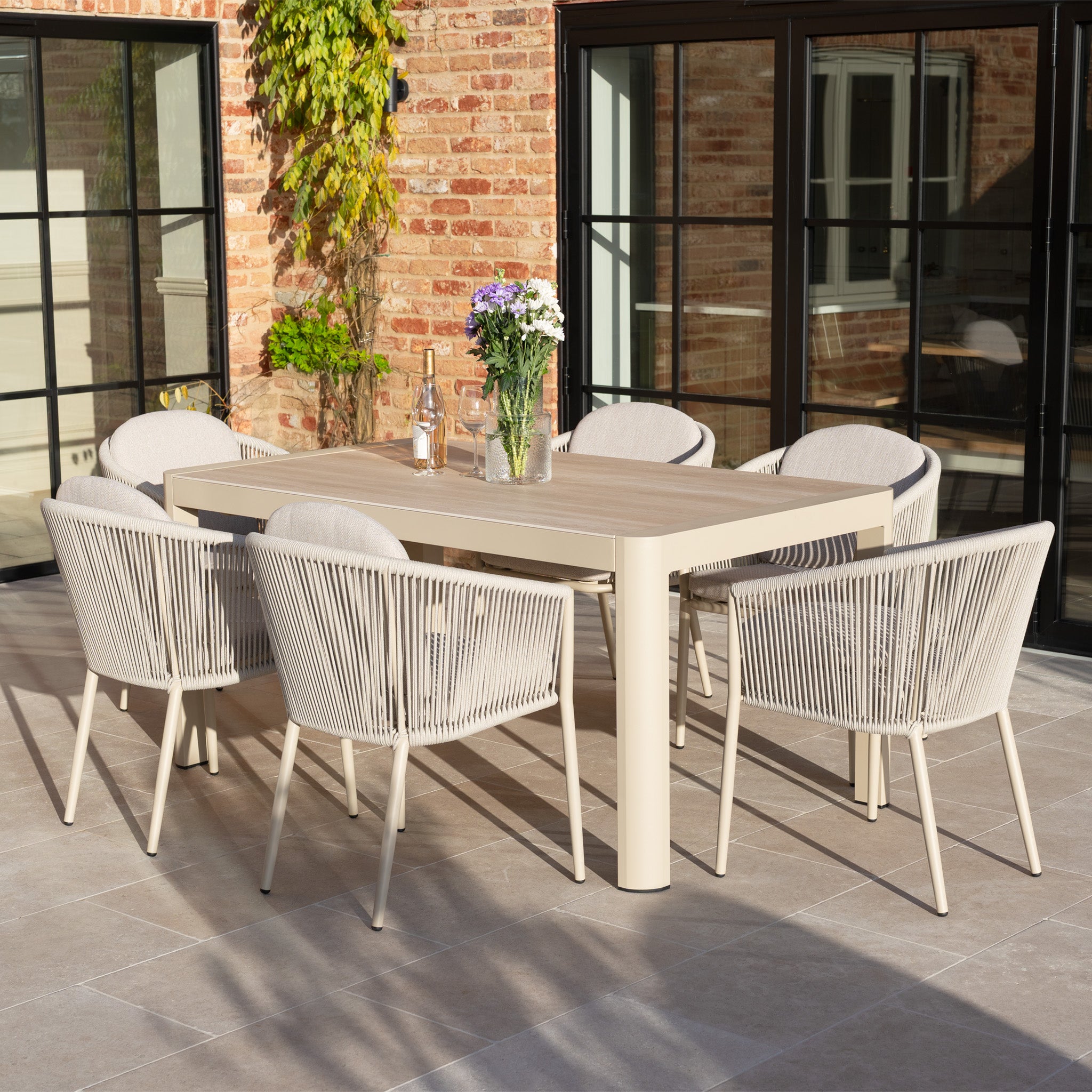 The Ravona 6 Seat Rope Extending Dining Set (162-222cm) in Latte features six beige chairs and a rectangular table, shown on a patio with glassware and flowers, and has an aluminium frame.