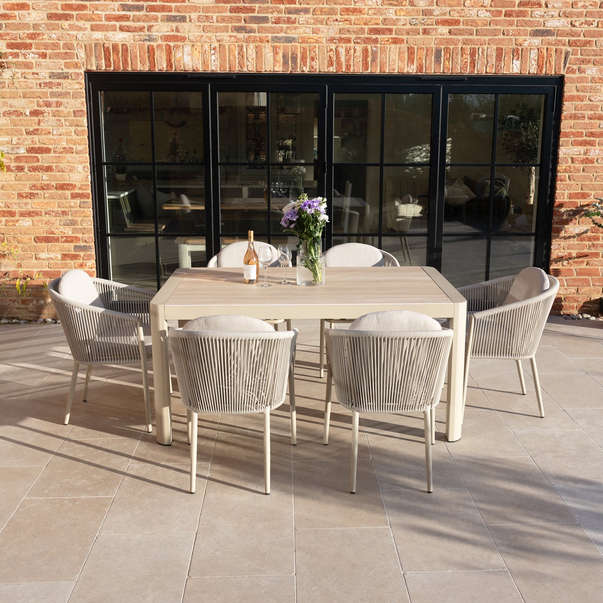 The Ravona 6 Seat Rope Extending Dining Set (162-222cm) in Latte includes six chairs and a rectangular aluminium table, stylishly placed on a patio by large glass doors, adorned with flowers and wine.
