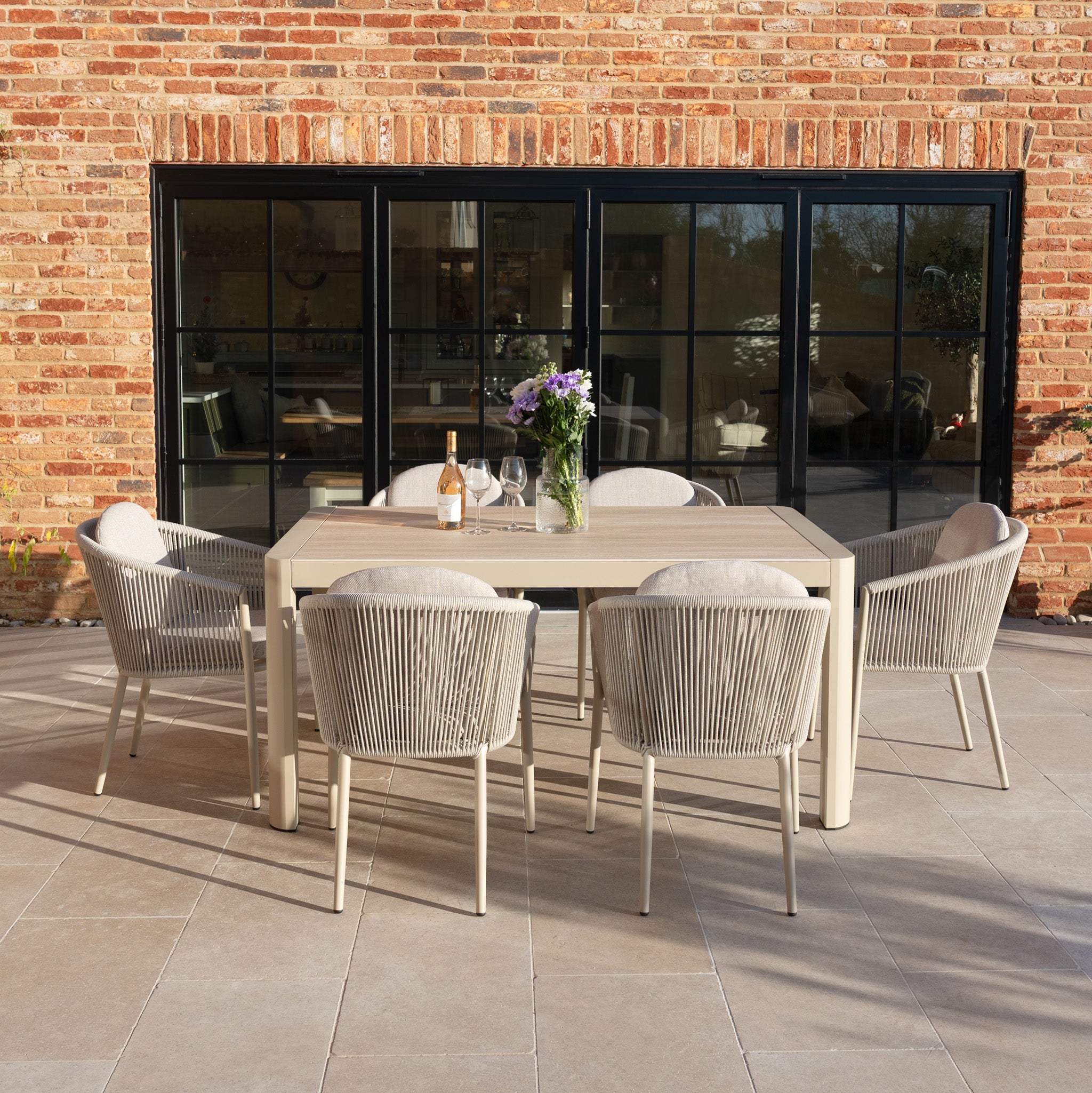 Enjoy sunny patio gatherings with the Ravona 6 Seat Rope Extending Dining Set (162-222cm) in Latte, featuring six chairs and a durable aluminium frame, perfect for outdoor dining by your charming brick house.