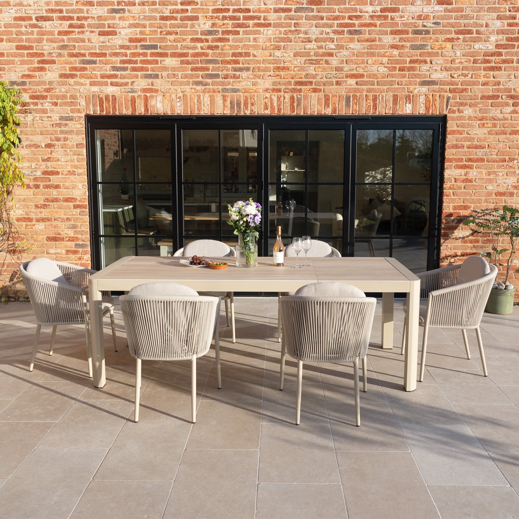 The Ravona 6 Seat Rope Extending Dining Set (162-222cm) in Latte includes six chairs and an aluminium-frame table, perfect for outdoor patio dining by a brick wall.