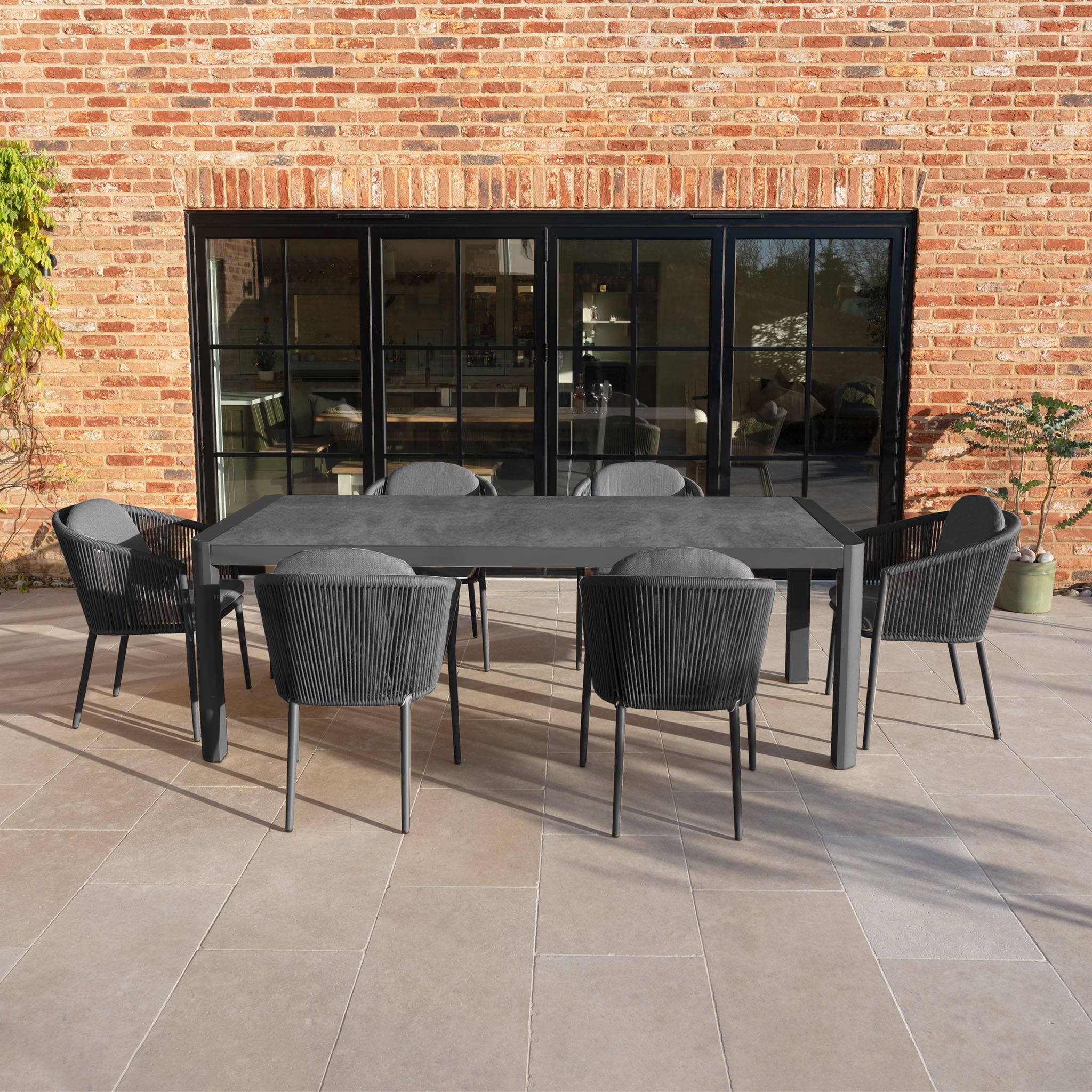 The Ravona 6 Seat Rope Extending Dining Set (162-222cm) in Charcoal features sleek black chairs and a roomy table, shown on a tiled patio with glass doors and a brick wall backdrop.