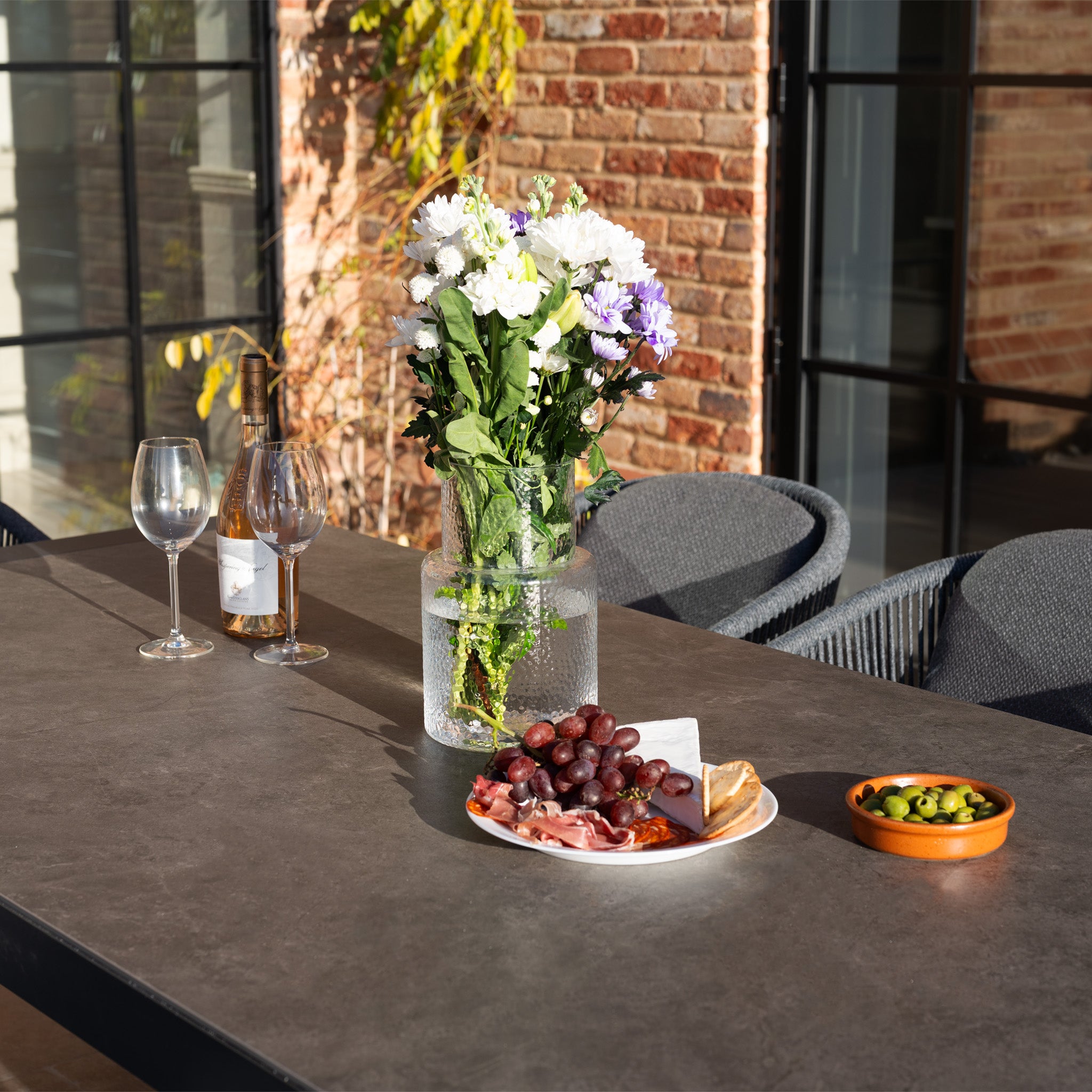 A vase of flowers, wine, glasses, snacks, and olives are beautifully arranged on the Ravona 8 Seat Rope Extending Dining Set (207-277cm) in Charcoal for stylish outdoor dining.