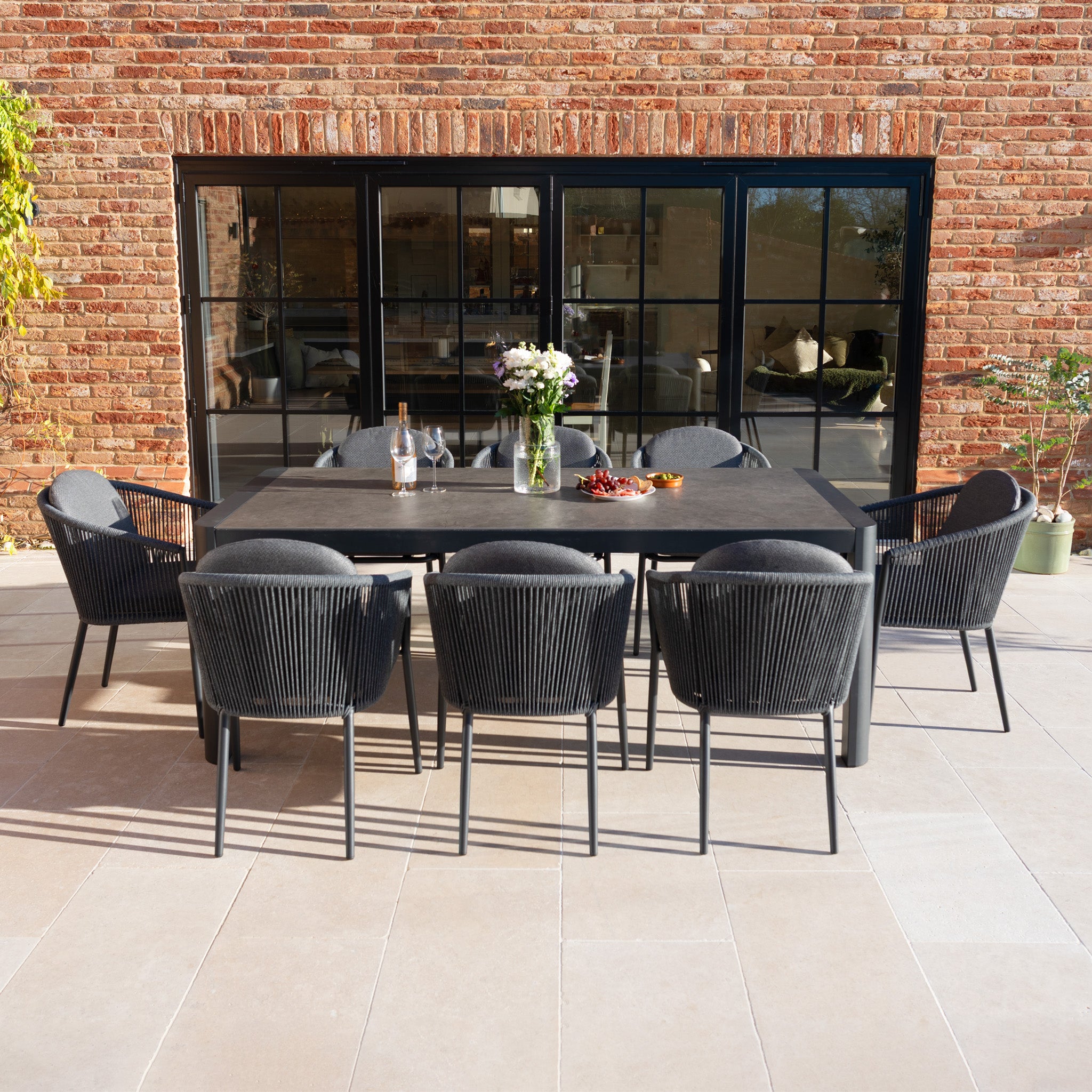The Ravona 8 Seat Rope Extending Dining Set (207-277cm) in Charcoal, with black chairs, is displayed on a patio with flowers and drinks before elegant glass doors.