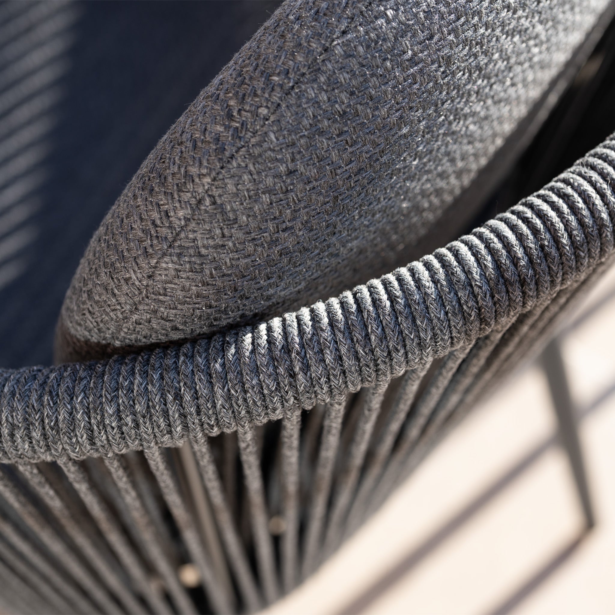 Close-up of a textured gray woven chair with fabric cushion and rope-wrapped frame, part of the Ravona 8 Seat Rope Extending Dining Set (207-277cm) in Charcoal, shown in sunlight.