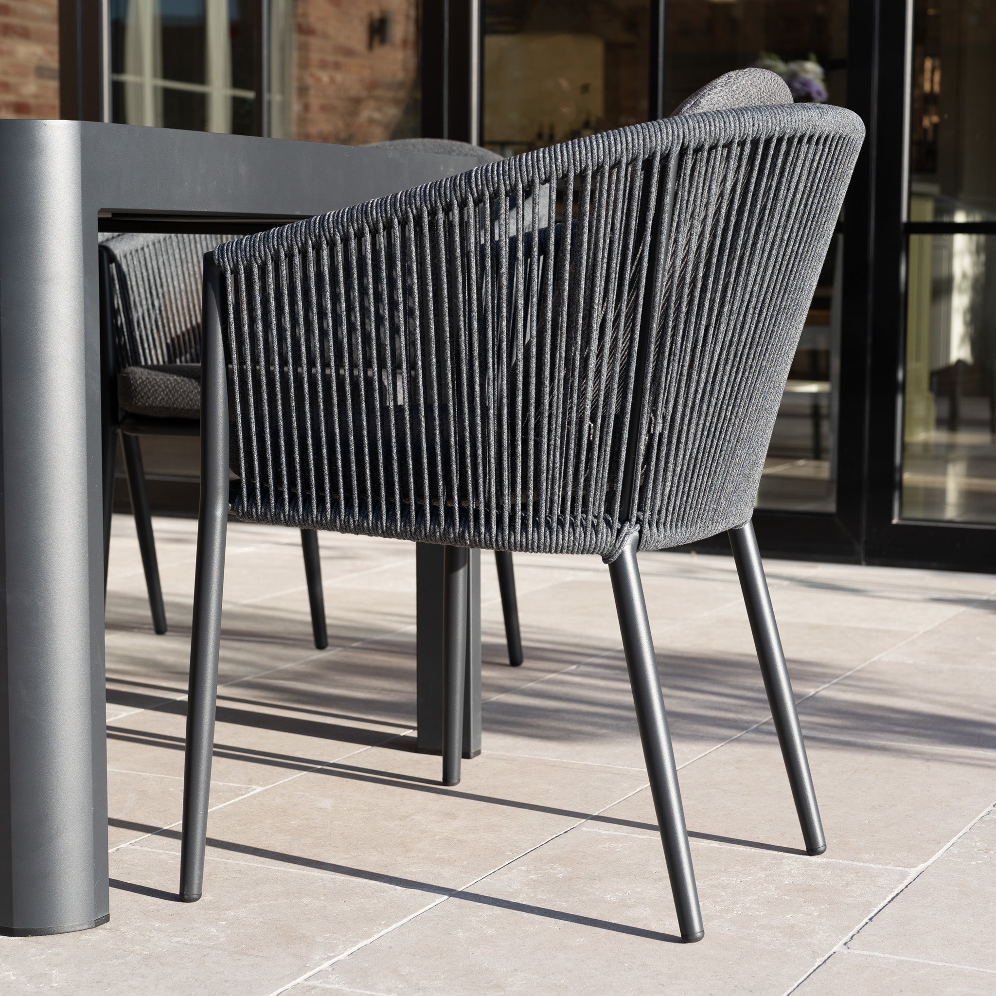 The Ravona 10 Seat Rope Extending Dining Set (207-277cm) in Charcoal features woven black chairs and a large table, shown on a tiled patio near glass doors.