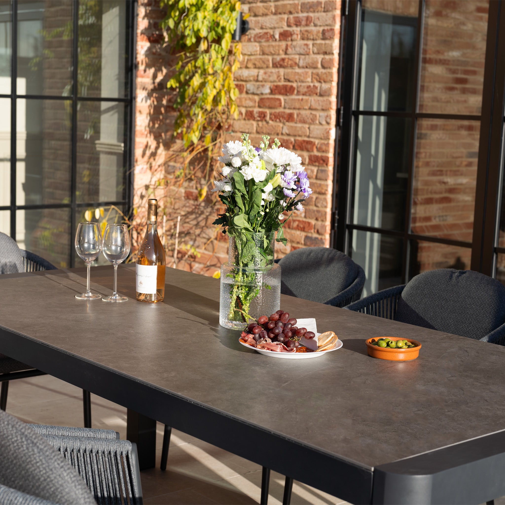 The Ravona 8 Seat Rope Extending Dining Set (207-277cm) in Charcoal is styled outdoors with wine, glasses, flowers, grapes, cheese, and olives arranged on the table against a brick building.