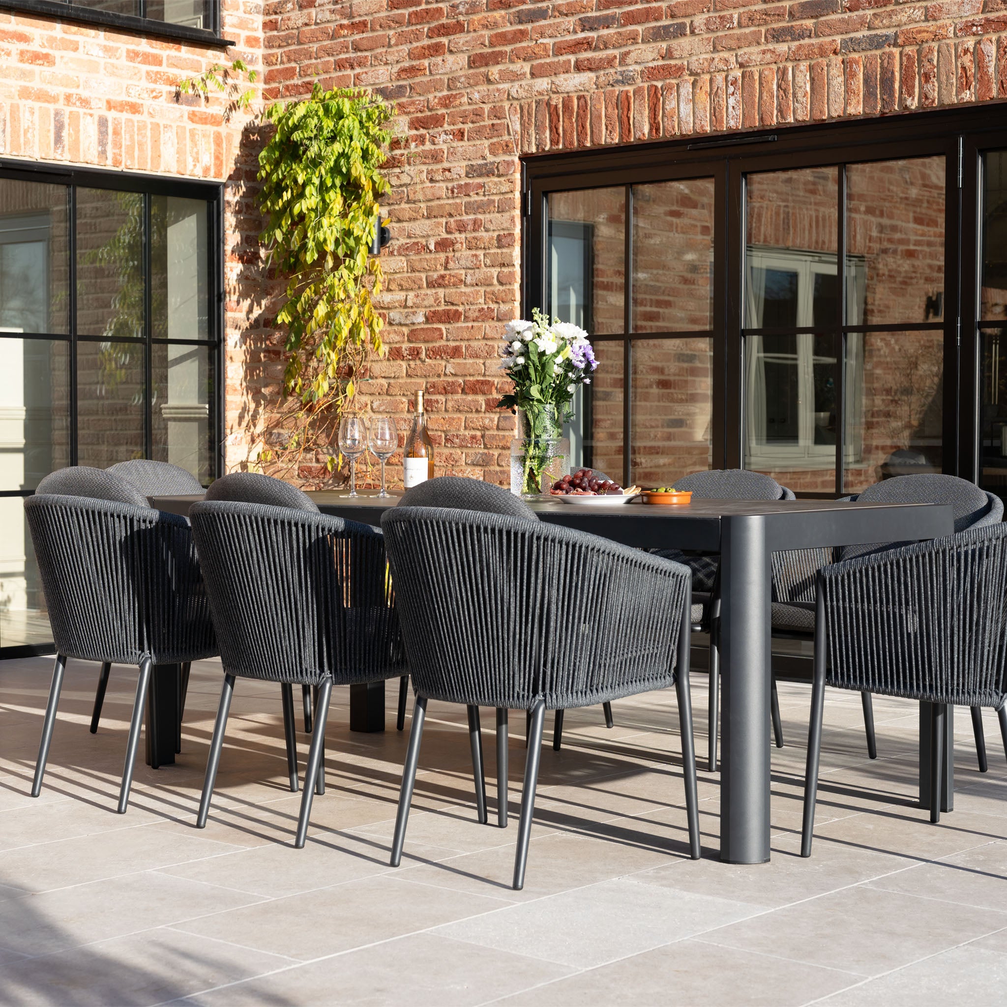Contemporary outdoor dining area featuring the Ravona 8 Seat Rope Extending Dining Set (207-277cm) in Charcoal with black chairs, a spacious table, flowers, and drinks next to a brick building.