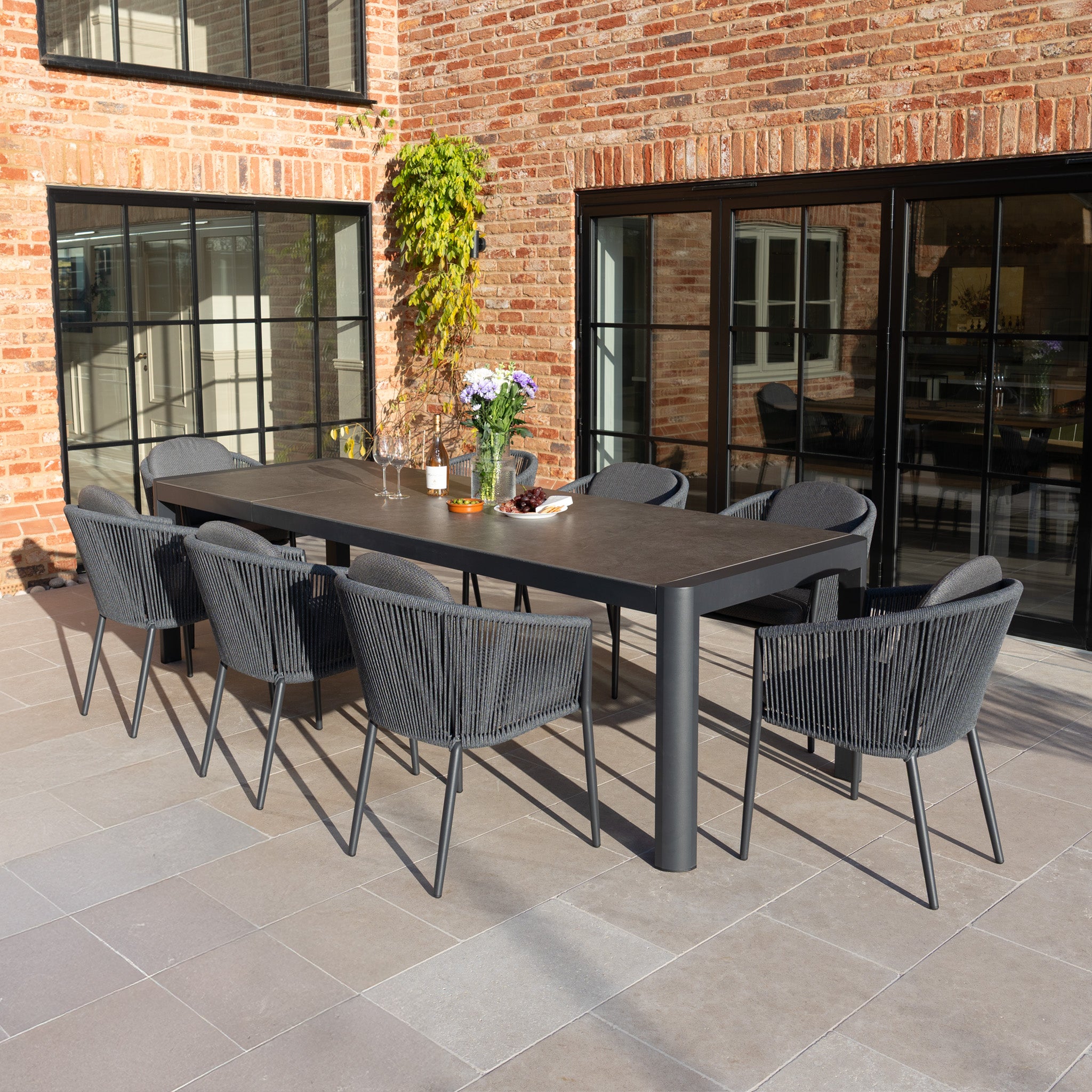 The Ravona 8 Seat Rope Extending Dining Set (207-277cm) in Charcoal features eight gray chairs, perfect for patio dining beside a brick house with large windows.