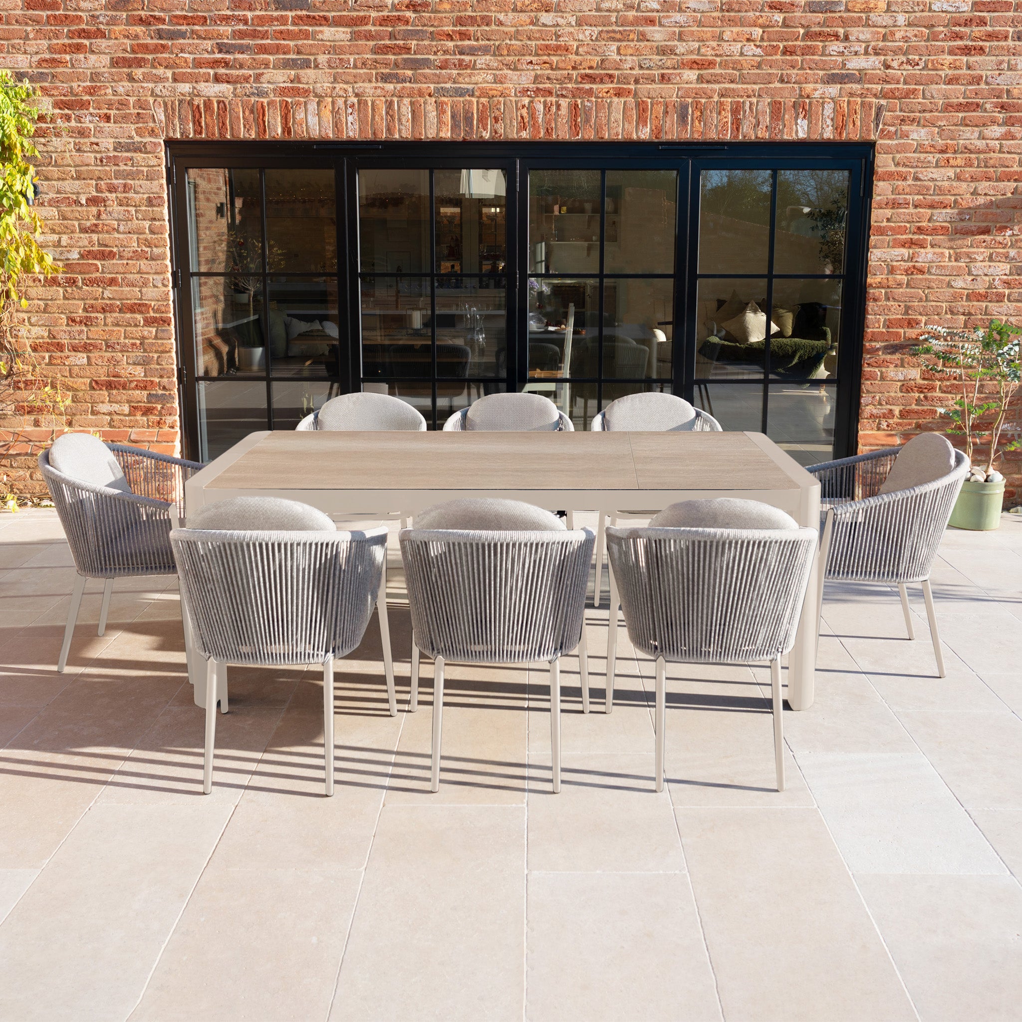 The Ravona 8 Seat Rope Extending Dining Set (207-277cm) in Latte features stylish chairs around an extendable table on a tiled patio, set against a brick wall with glass doors.