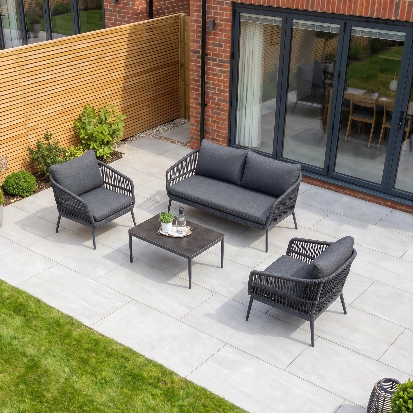 A modern patio featuring the Rhea 2 Seat Sofa Set in Grey, complemented by a matching coffee table, lush plants, glass doors, and a wooden fence for a stylish outdoor look.