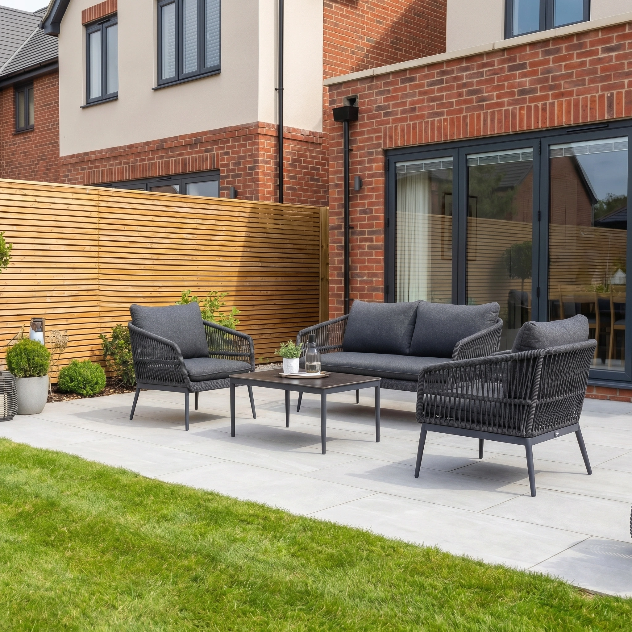 Modern patio with sleek aluminum garden furniture, featuring the Rhea 2 Seat Sofa Set in Grey, a table, and potted plants beside a brick house bordered by a wooden fence.
