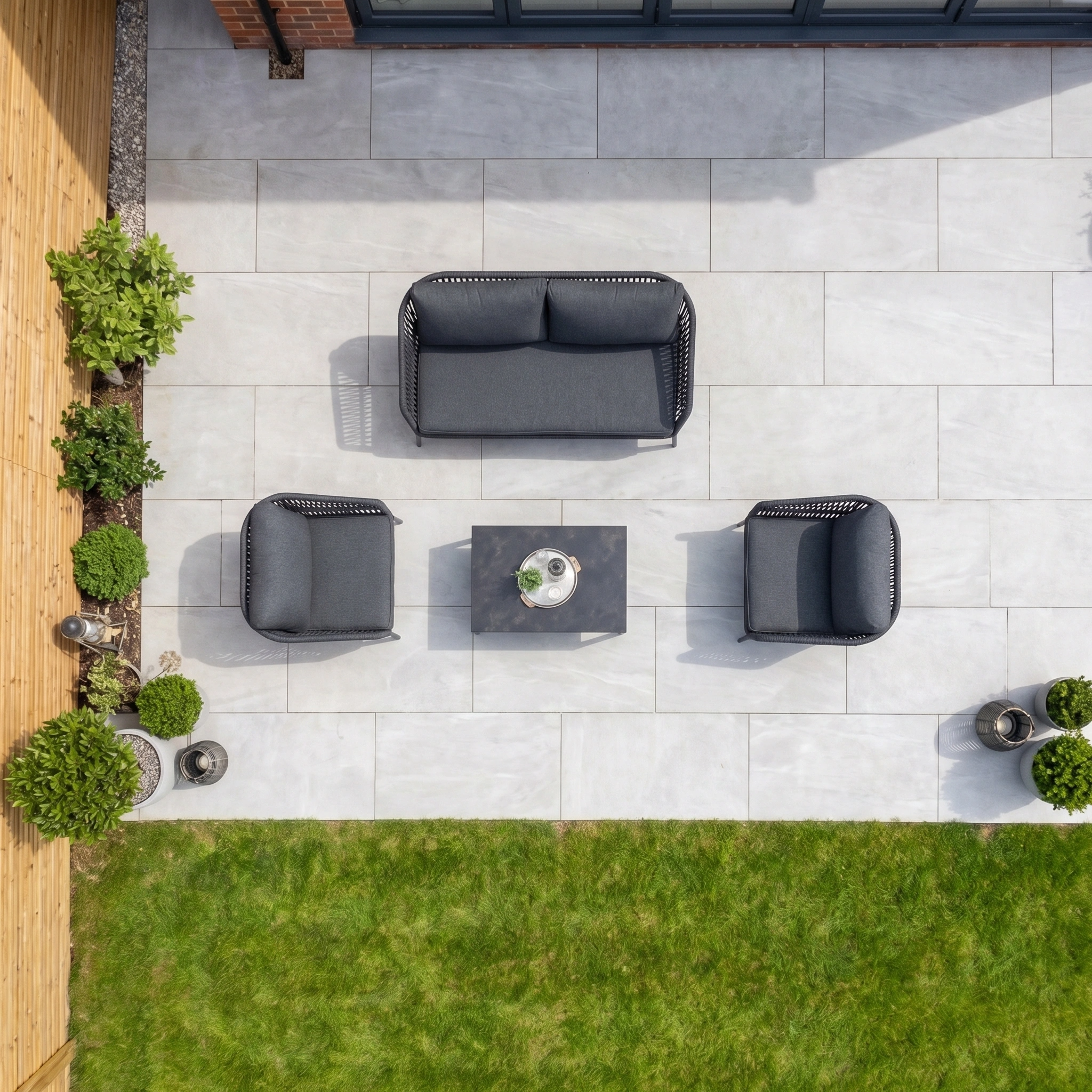 Modern patio with gray tiles, a sleek Rhea 2 Seat Sofa Set in Grey paired with black outdoor seating, a small table, green plants, and a lawn below.