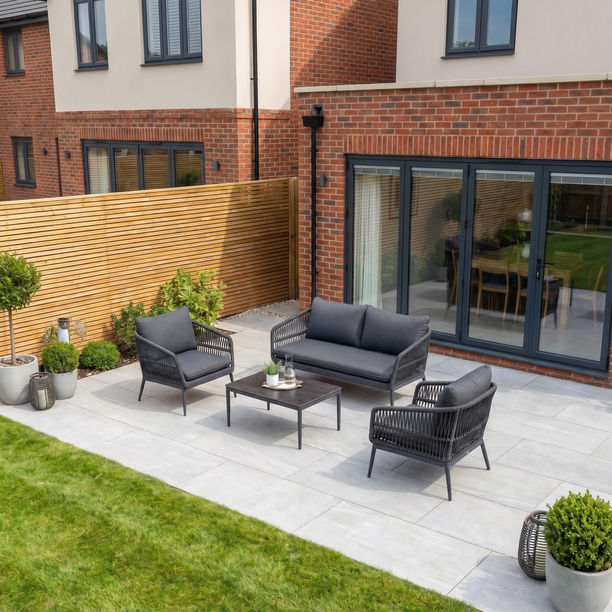 Modern patio featuring the Rhea 2 Seat Sofa Set in Grey, potted plants, and sliding glass doors beside a lawn and brick house.