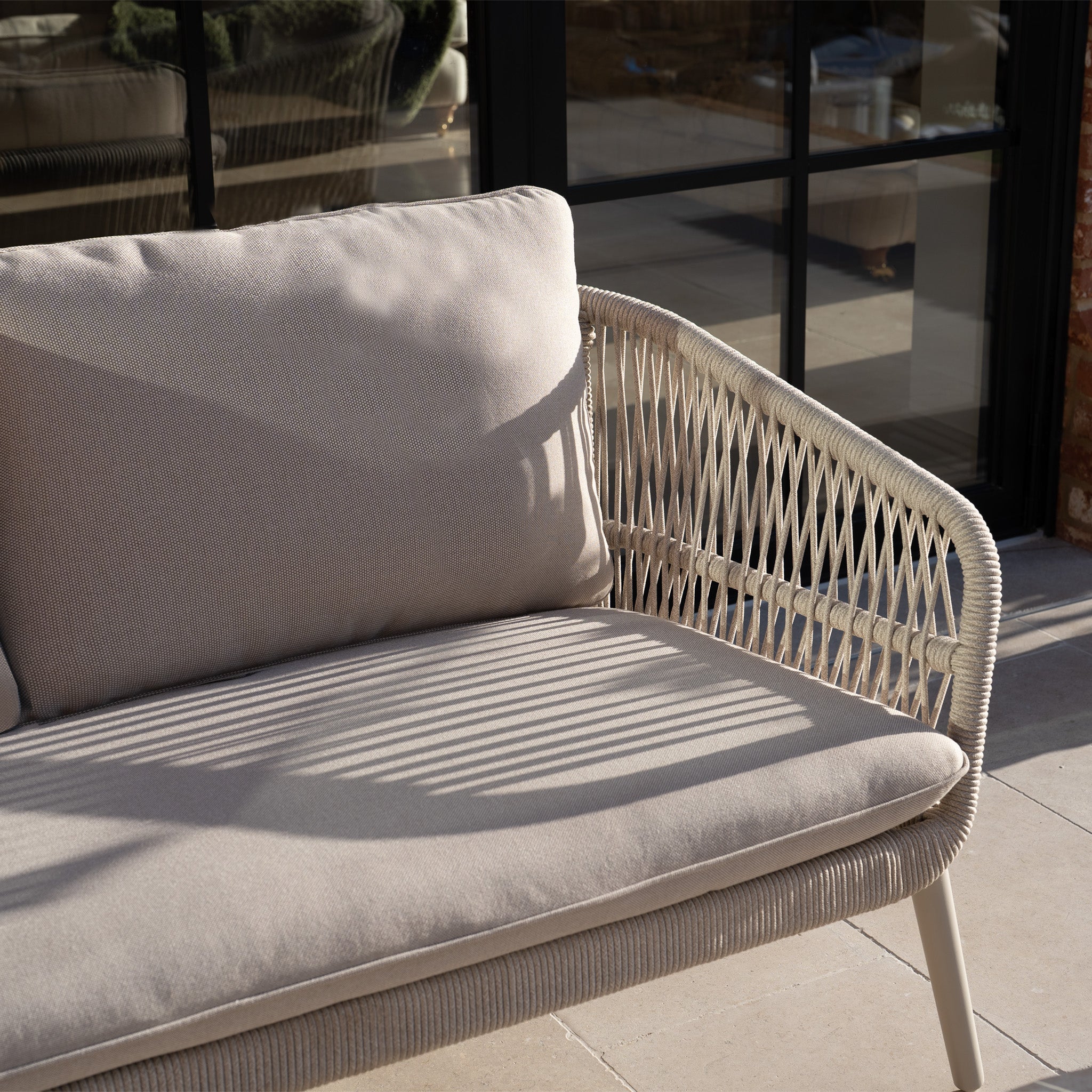 The Rhea 2 Seat Sofa Set in Latte features beige wicker, comfy cushions, and a durable aluminum frame, with sunlight creating windowpane shadows across the seat and backrest.