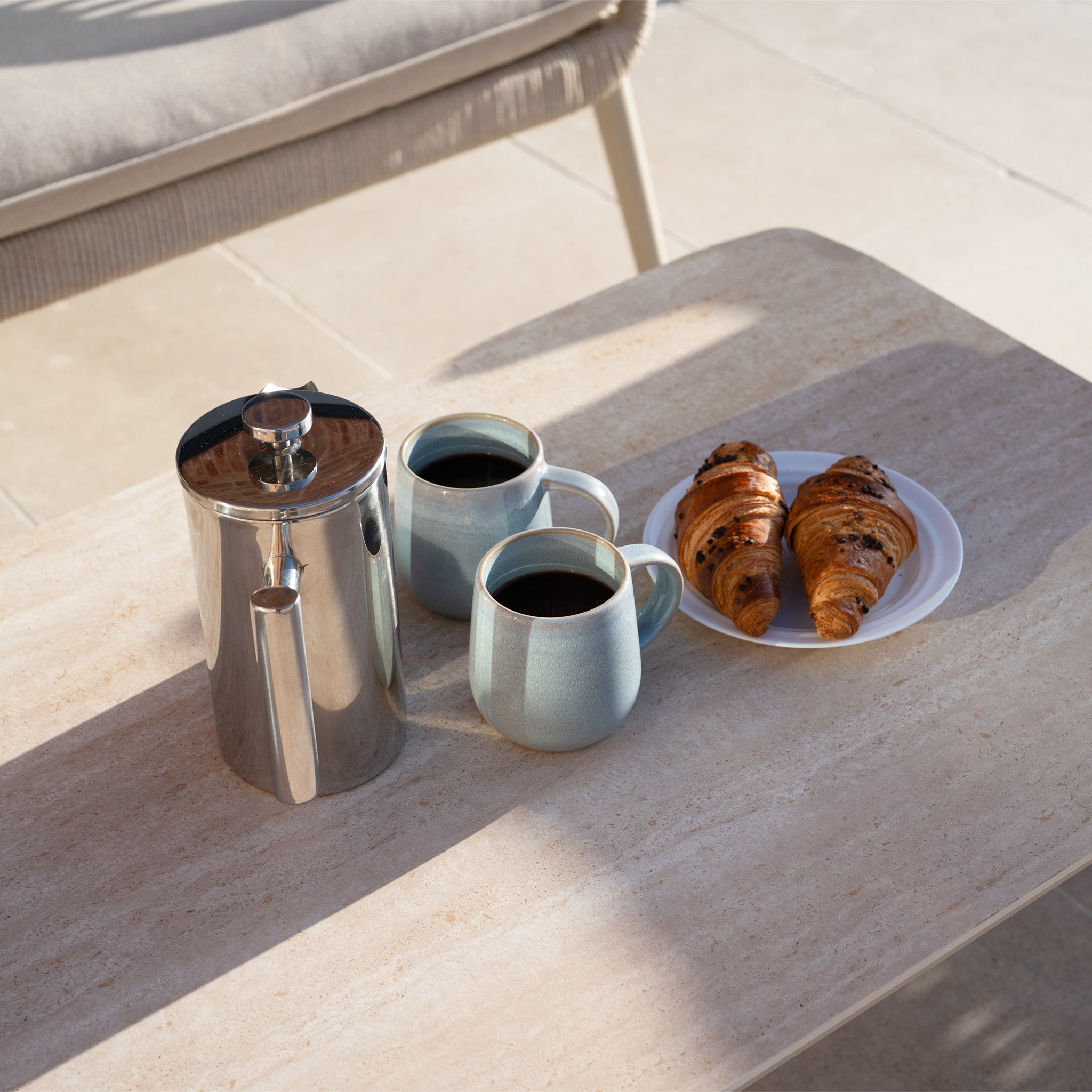 A coffee pot, two mugs, and two croissants rest on a sunlit table beside stylish aluminium garden furniture and the cozy Rhea 2 Seat Sofa Set in Latte.