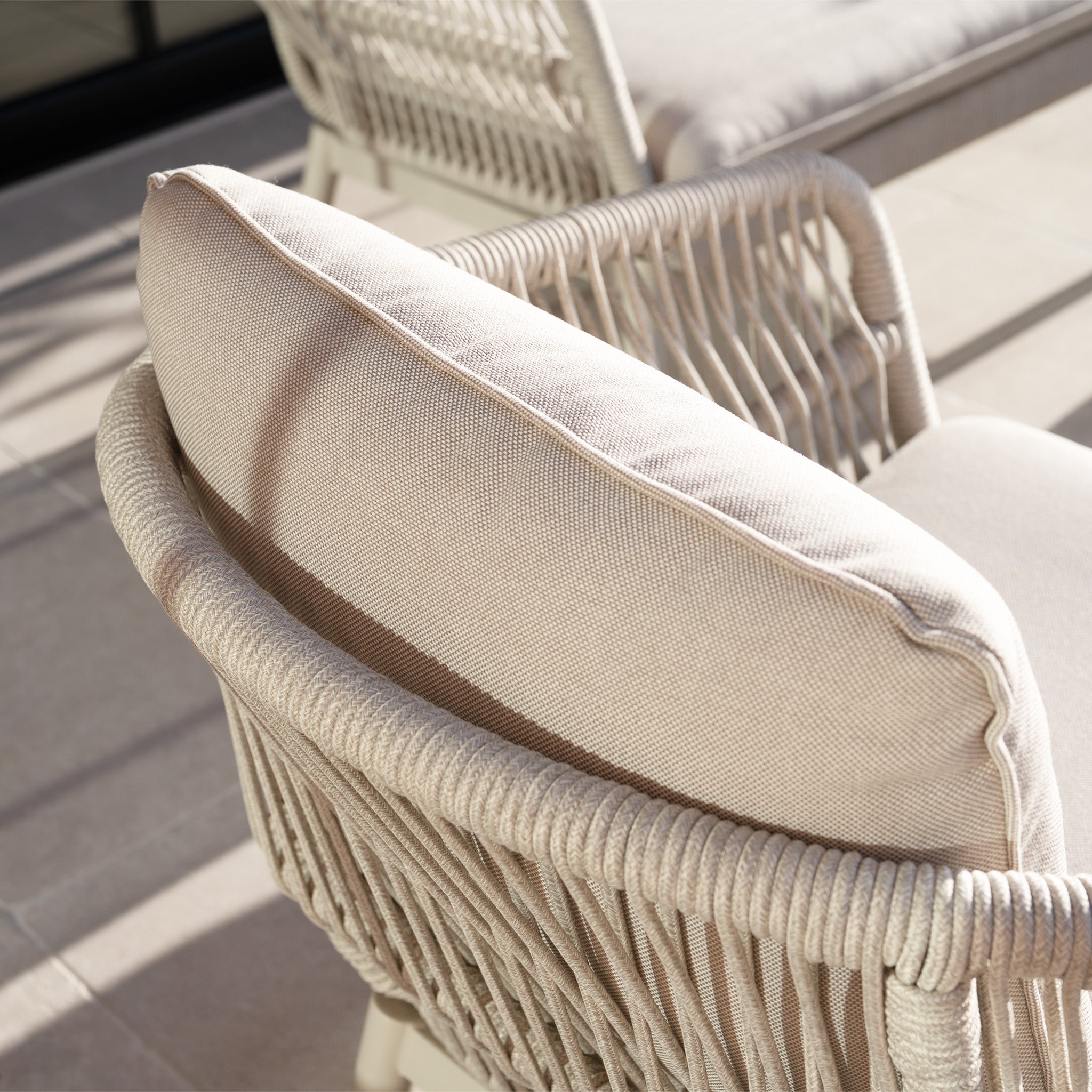 The Rhea 2 Seat Sofa Set in Latte features a beige woven design with thick cushions, shown on a tiled patio in sunlight—ideal for enhancing your aluminium garden furniture collection with style and comfort.
