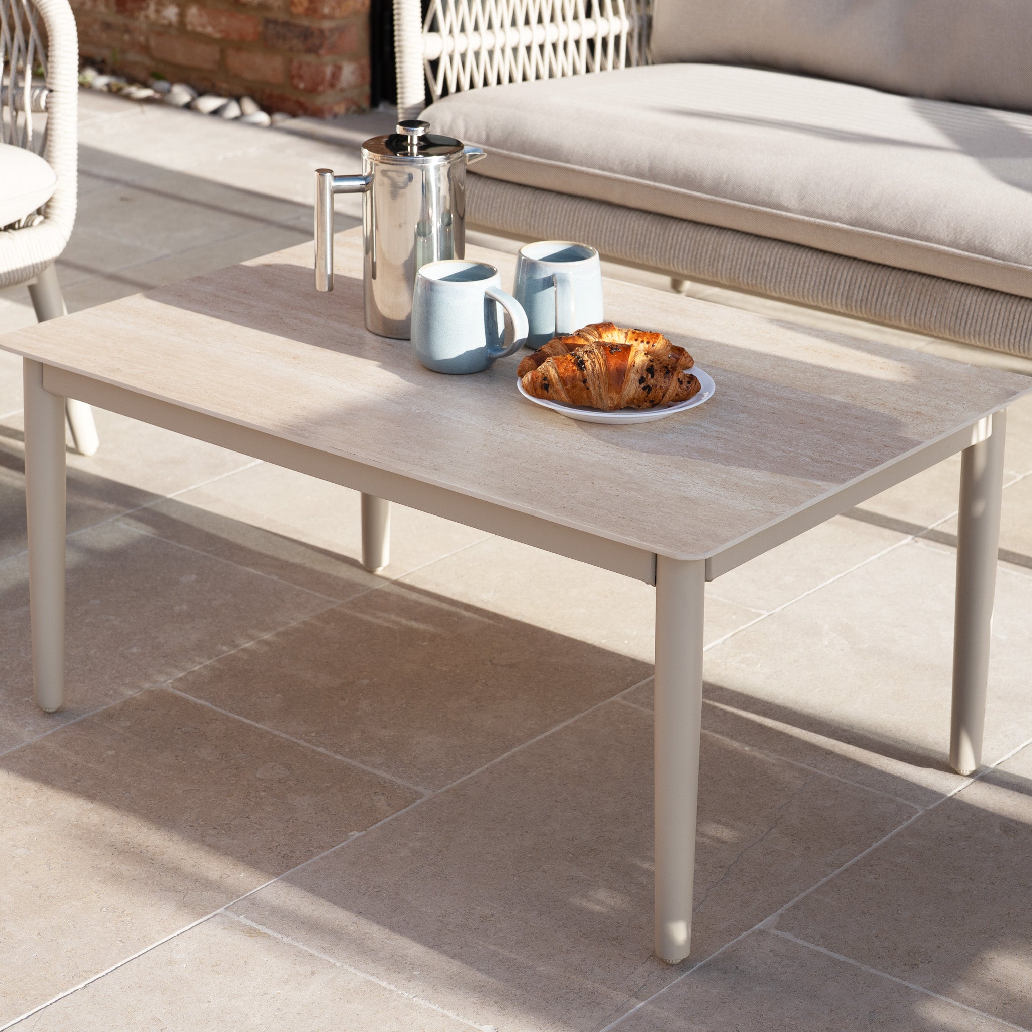 Outdoor coffee table with a coffee pot, two mugs, and a plate of croissants on a sunny patio next to the Rhea 2 Seat Sofa Set in Latte.