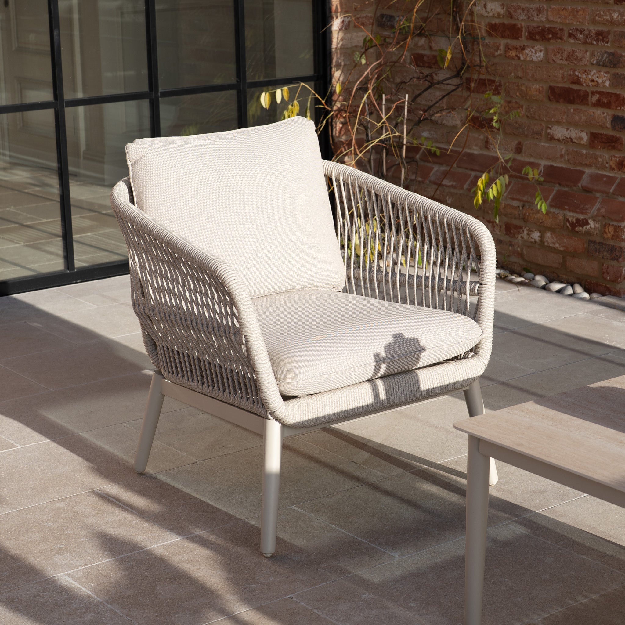 The Rhea 2 Seat Sofa Set in Latte, featuring woven sides and soft cushions, is part of a chic aluminium garden furniture collection and sits on a tiled patio near a brick wall.