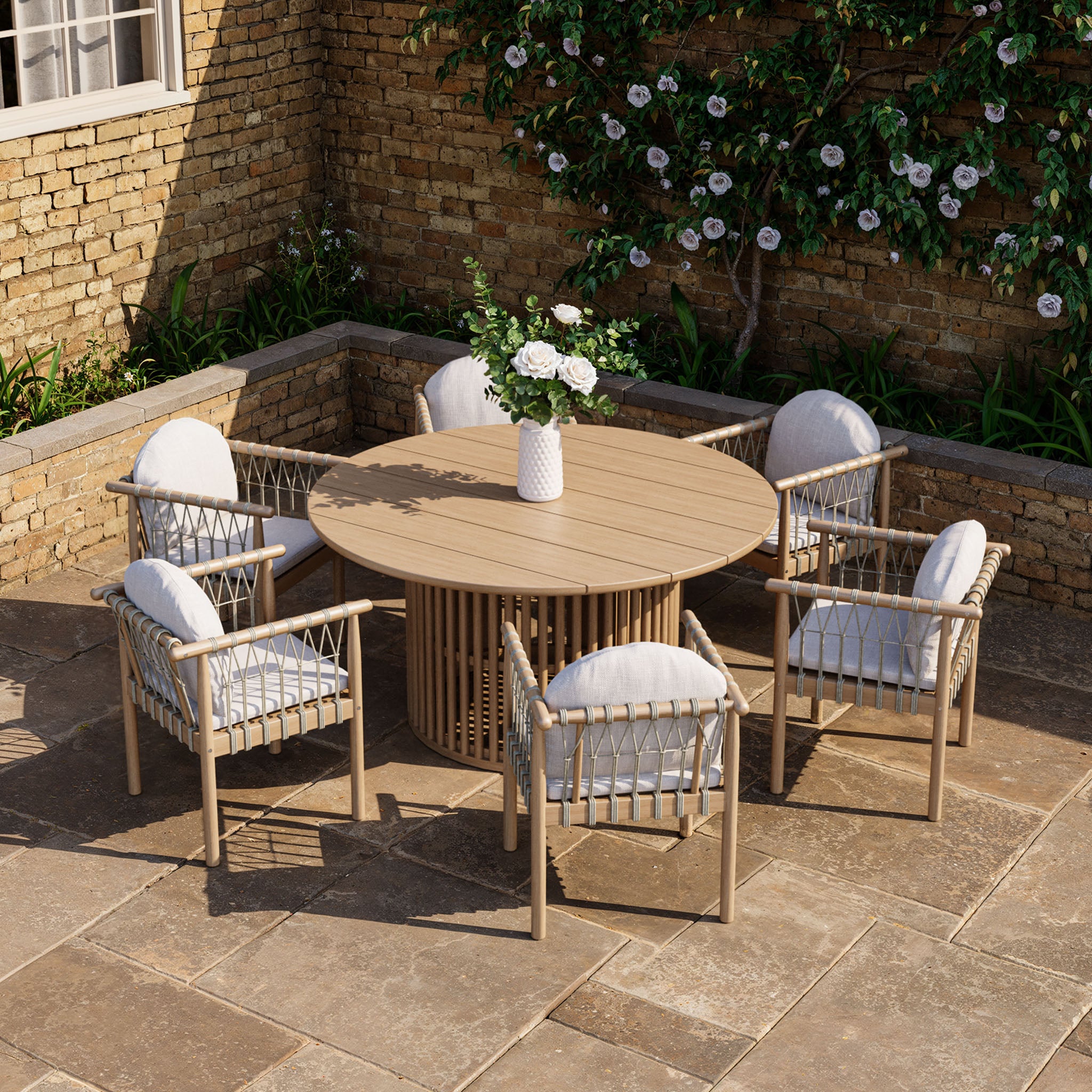 The Paia 6 Seat Round Dining Set in Linen includes a sustainably sourced Acacia wood table and six water-resistant polyester cushioned chairs, elegantly set against a brick wall with a vase of fresh flowers.