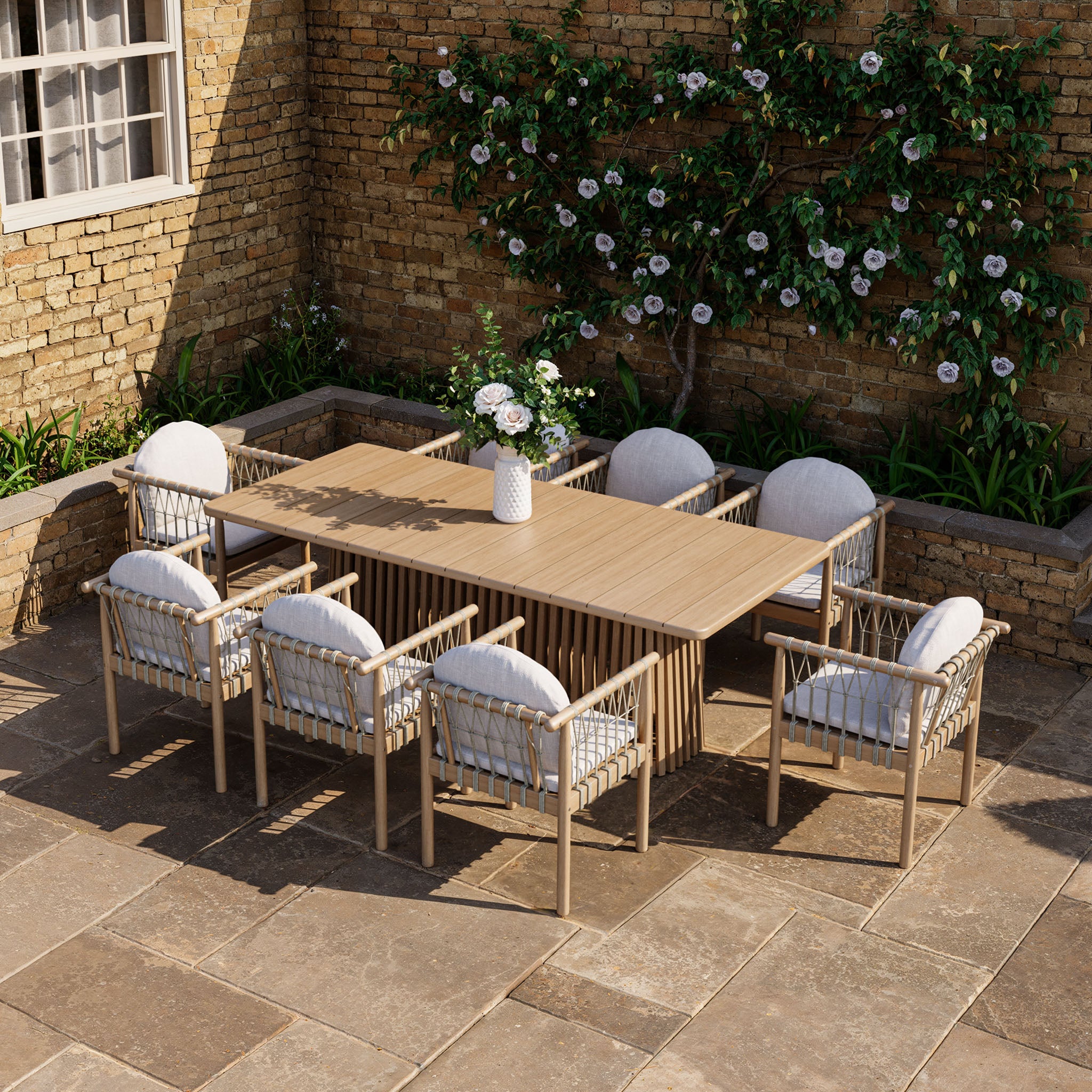 The Paia 8 Seat Rectangular Dining Set in Linen offers a stylish outdoor dining experience with sustainably sourced Acacia wood and plush white cushions, ideal for patio spaces against charming brick walls with blooming plants.