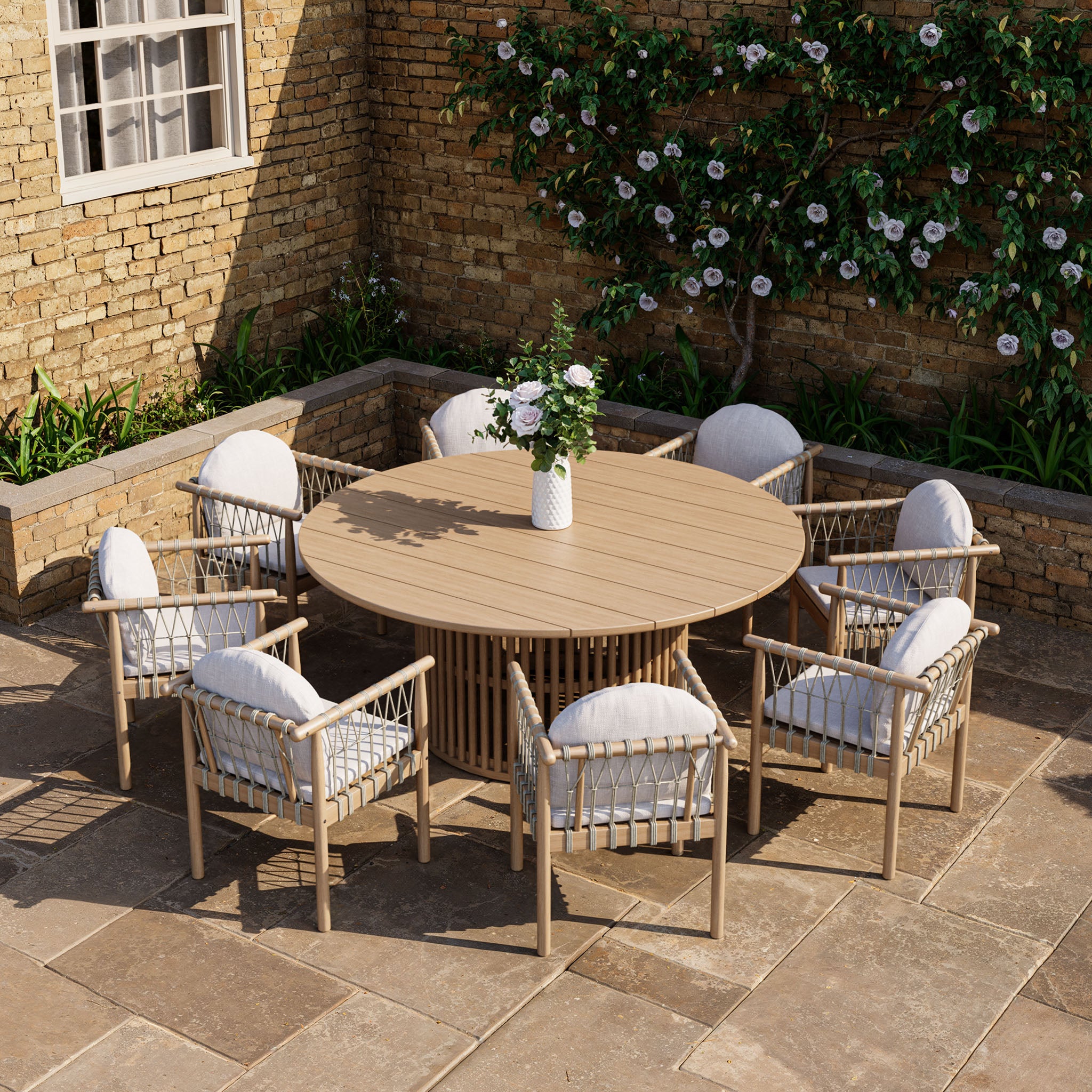 The Paia 8 Seat Round Dining Set in Linen, made from sturdy acacia wood, includes cushioned chairs around a round table. Situated on a stone patio by a flowery brick wall, it creates an inviting outdoor dining area.