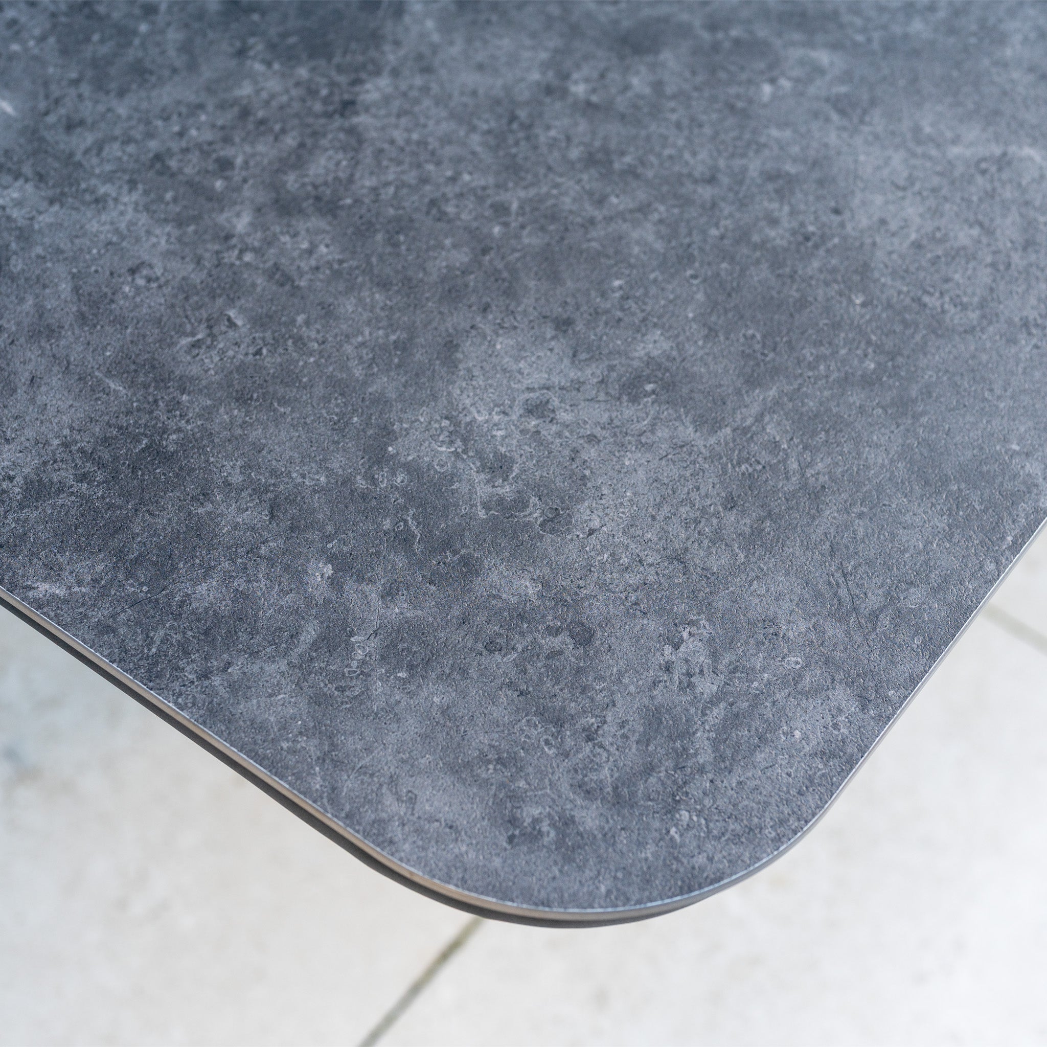 Close-up of the dark gray, stone-textured tabletop with a rounded corner above tiled flooring—ideal for pairing with the Savona Luxury Outdoor Large Corner Sofa Set with Altura Rising Table in Truffle.