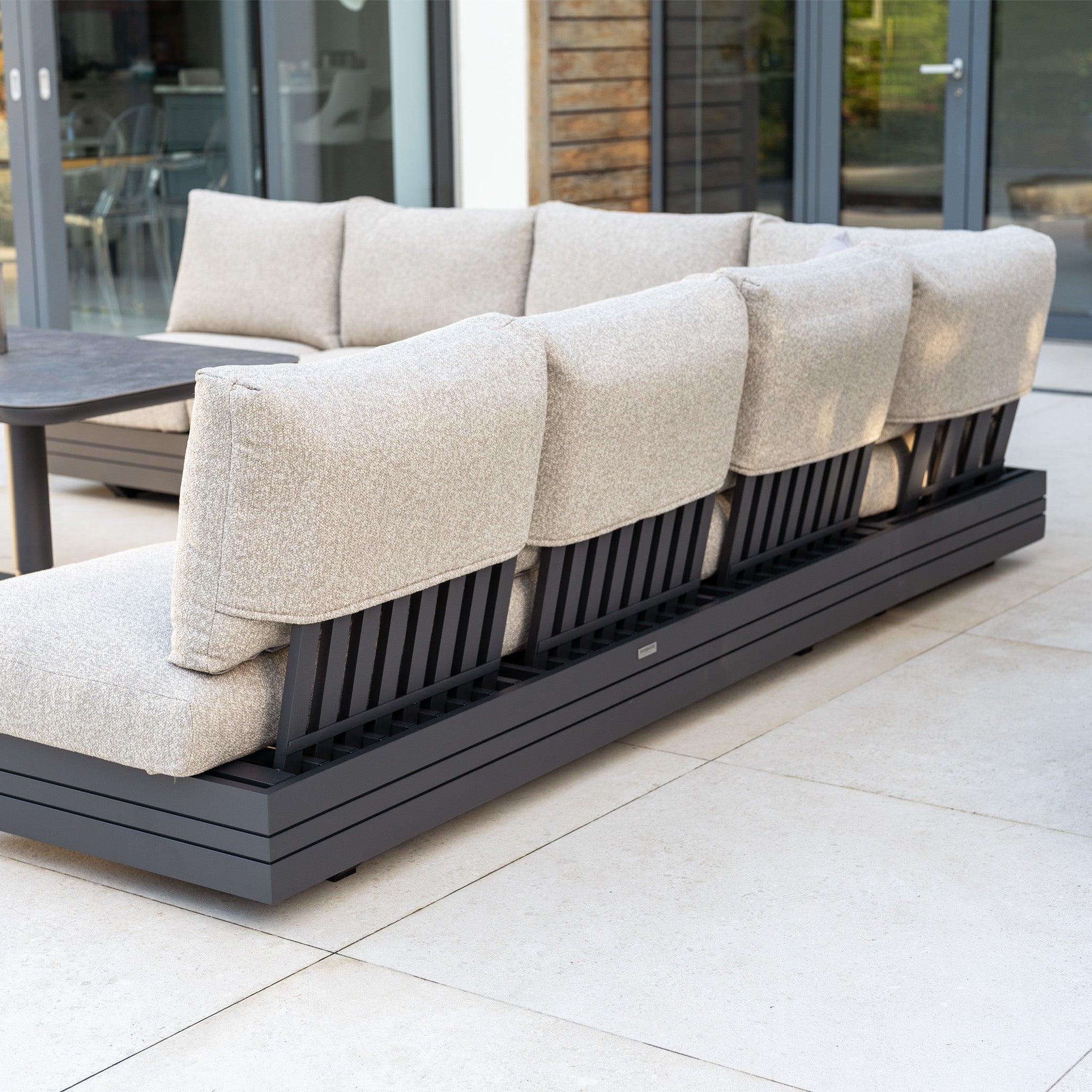 The Savona Luxury Outdoor Large Corner Sofa Set with Altura Rising Table in Truffle sits on a tiled patio near glass doors, its beige modular seating and black frame paired with a sleek rising table.