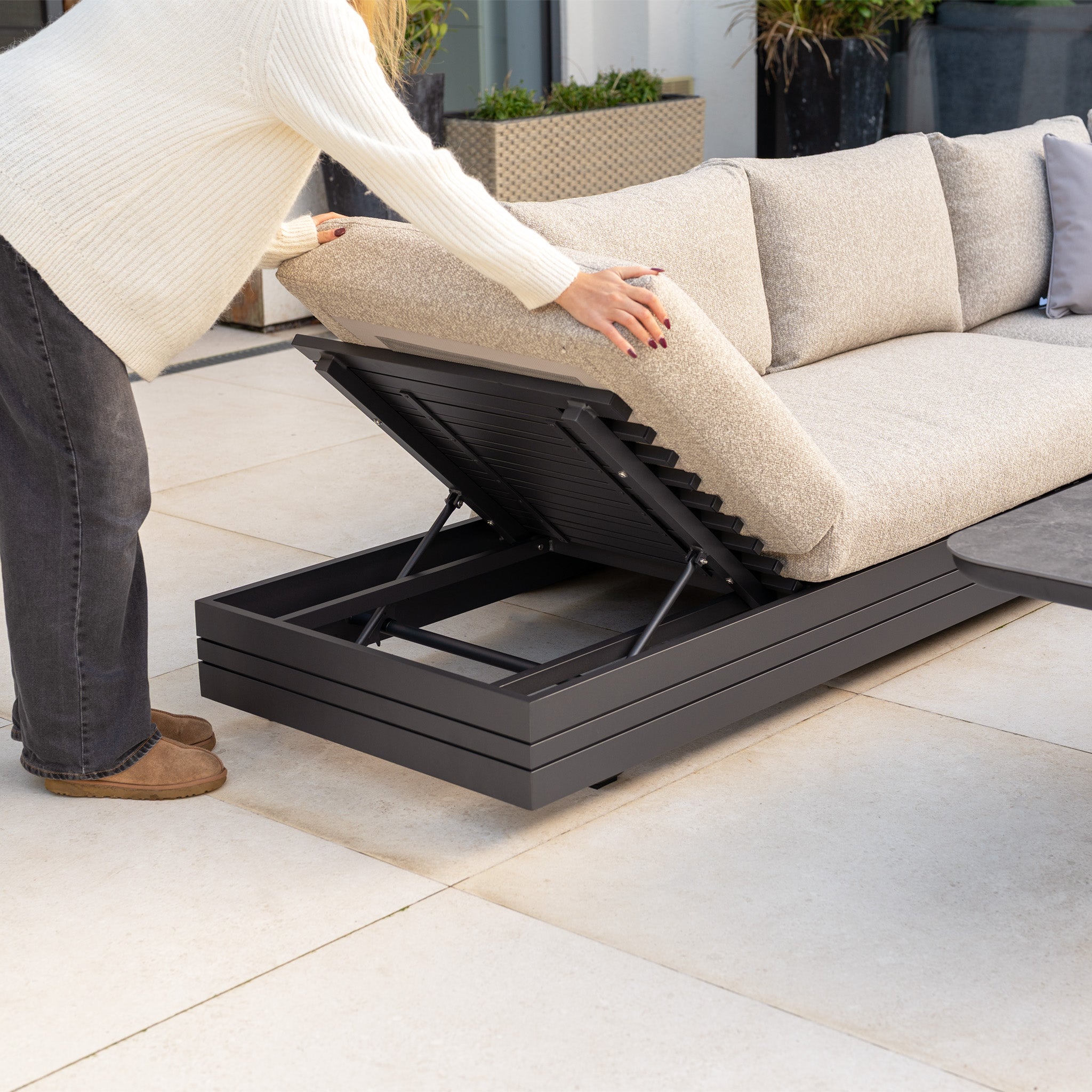 A person lifts the cushioned seat of the Savona Luxury Outdoor Large Corner Sofa Set with Altura Rising Table in Truffle to reveal hidden storage underneath.