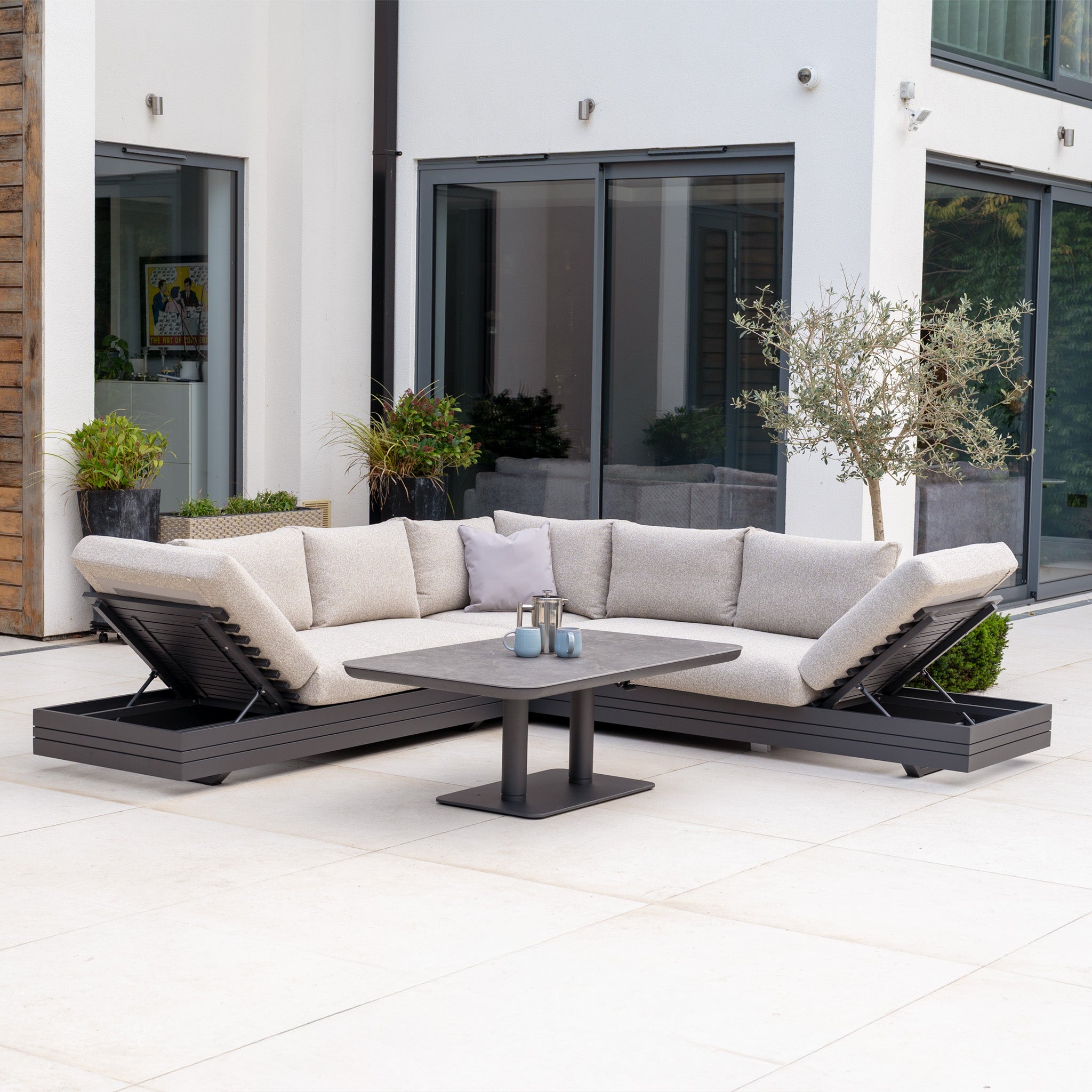 Modern outdoor patio featuring the Savona Luxury Outdoor Large Corner Sofa Set with beige cushions and an Altura Rising Table in Truffle, arranged by large glass doors and accented with potted plants.