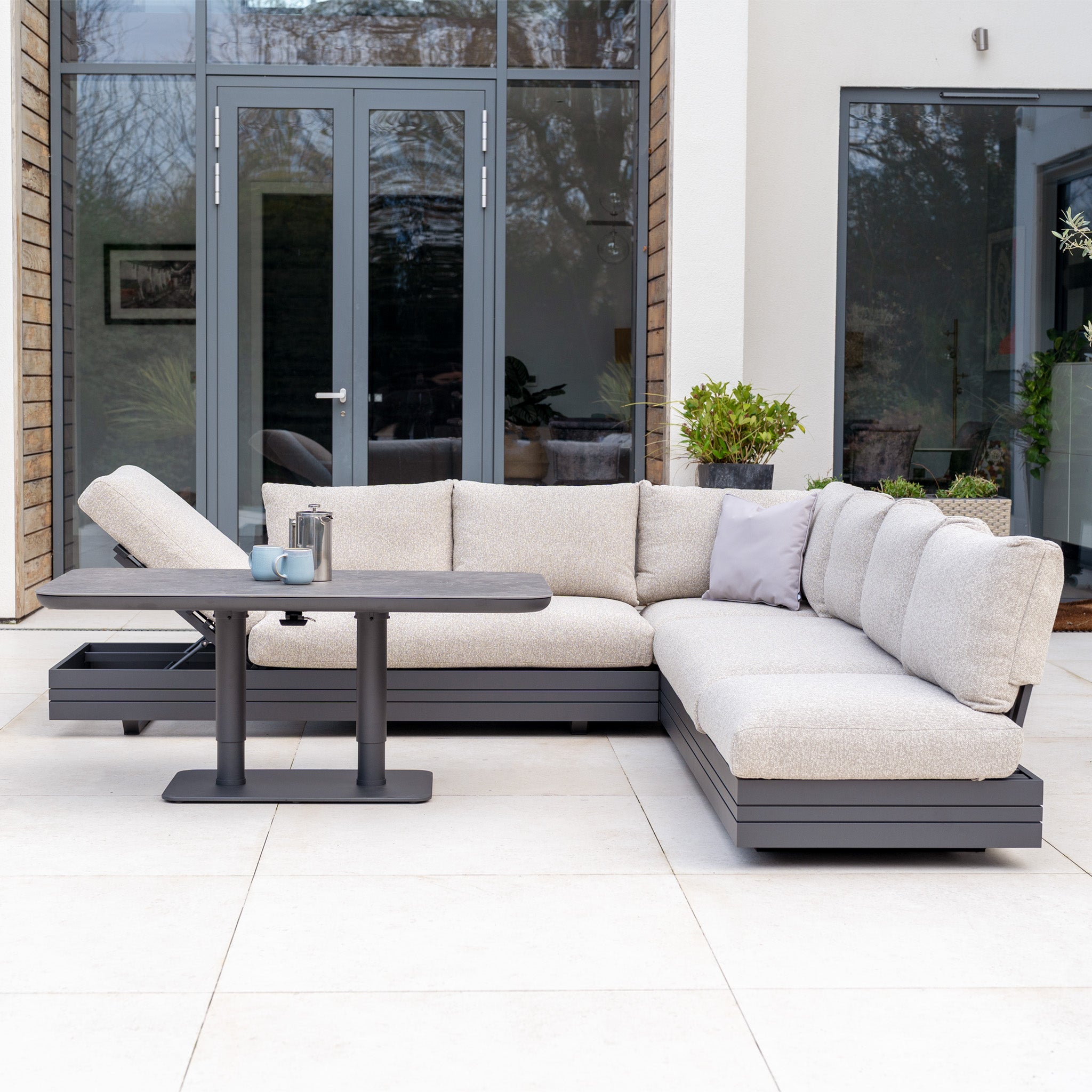 Modern outdoor patio featuring the Savona Luxury Outdoor Large Corner Sofa Set in Truffle, arranged in an L-shape, paired with the Altura Rising Table and glass doors in the background.