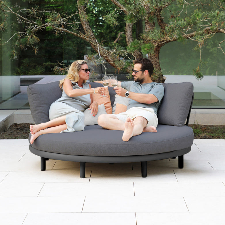 A man and woman relax on a Selene Daybed in Charcoal, smiling at each other and clinking wine glasses as they enjoy the comfort of this luxury patio furniture.