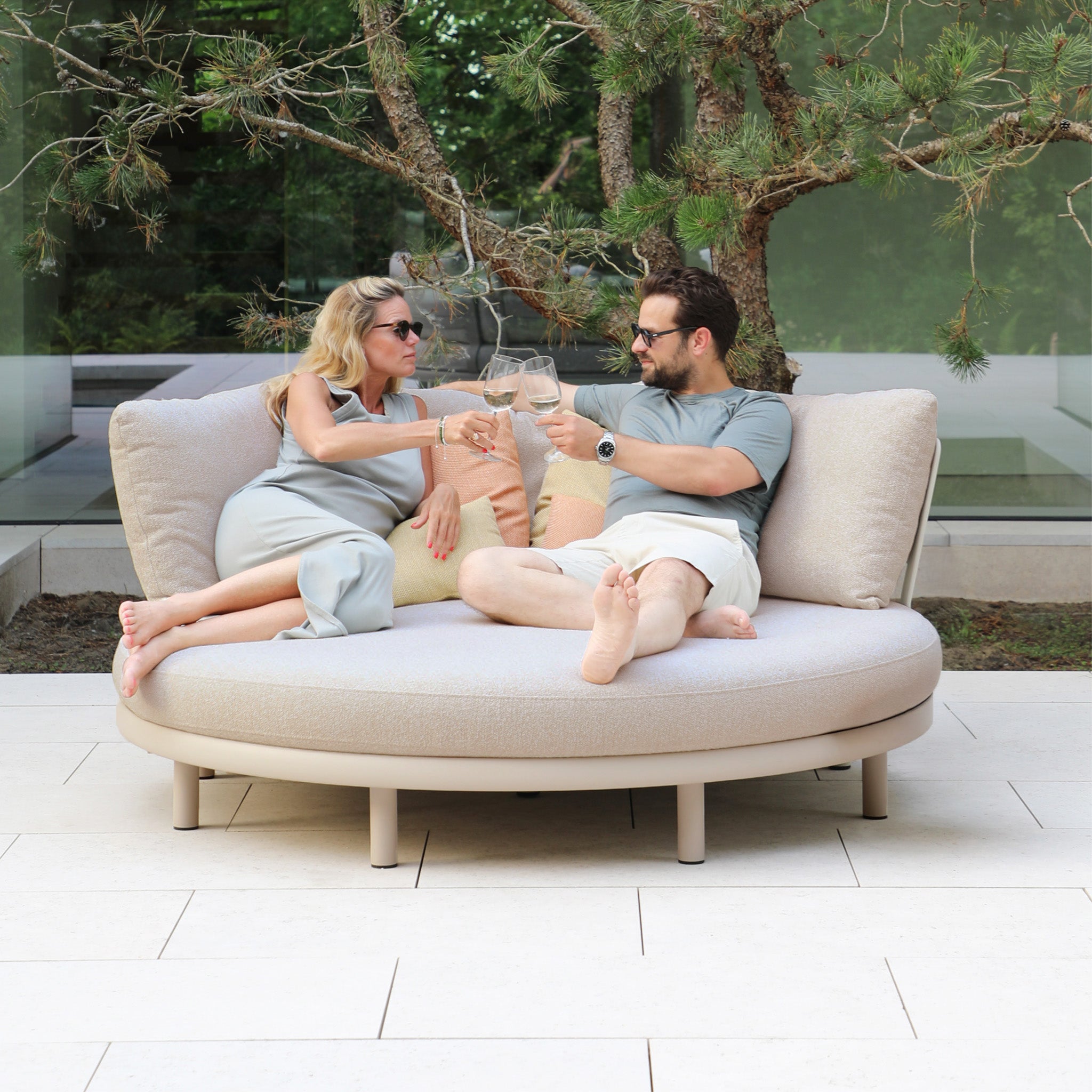 A man and woman relax on the Selene Daybed in Fawn, clinking wine glasses as they unwind outdoors with lush greenery in the background.