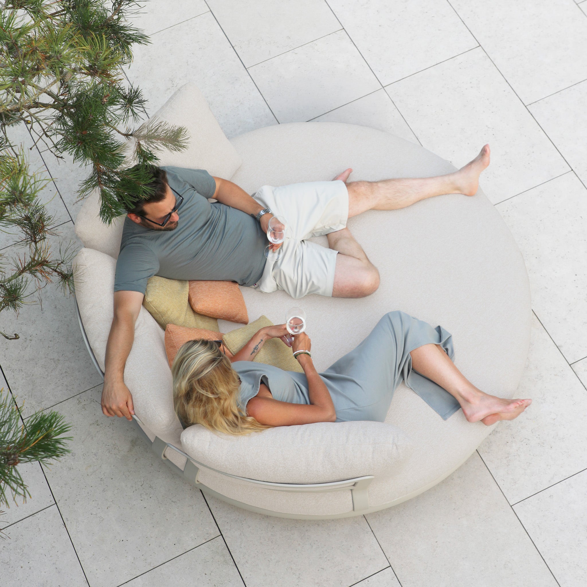 A man and woman relax on the Selene Daybed in Fawn, holding drinks and chatting, as seen from above.