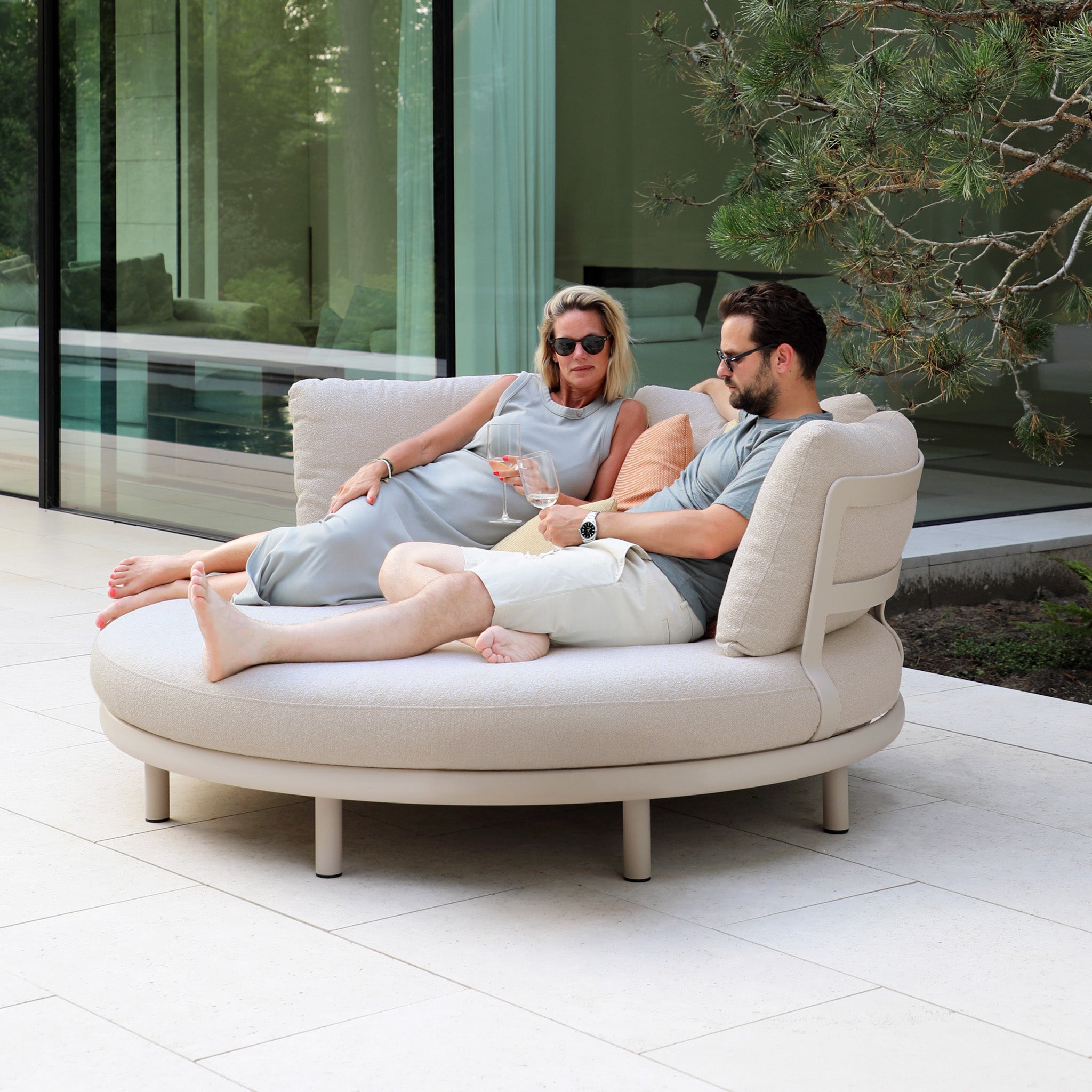 Two people unwind on the Selene Daybed in Fawn, sipping drinks and enjoying comfort near a modern glass building surrounded by lush greenery.