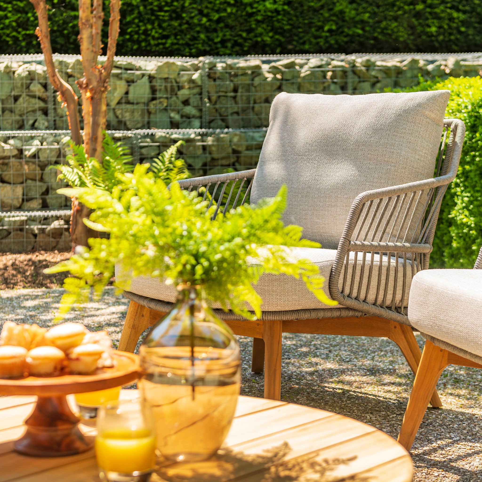 Outdoor patio featuring Sempre furniture, including a 2-seat sofa set with a teak louvred table and footstool in light grey. Juice and pastries rest on the table, while potted ferns accent the space with a green hedge in the background. Each piece is handmade with passion for your comfort.