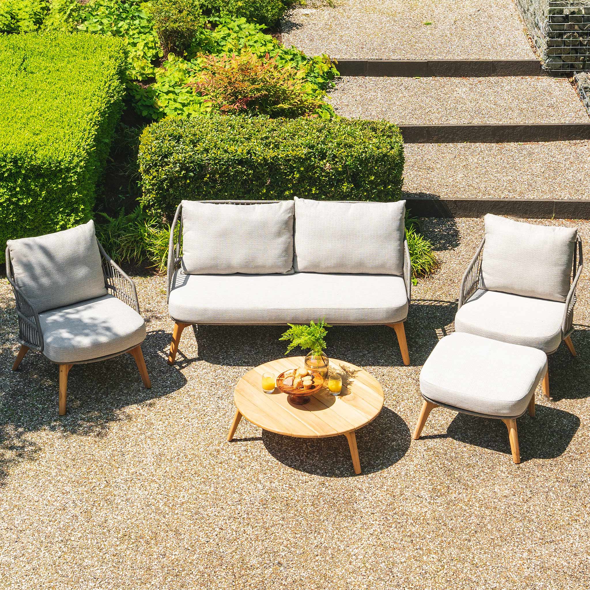 Outdoor seating area with the Sempre 2 Seat Sofa Set in light grey cushioned furniture, a teak louvred coffee table, and greenery in the background.