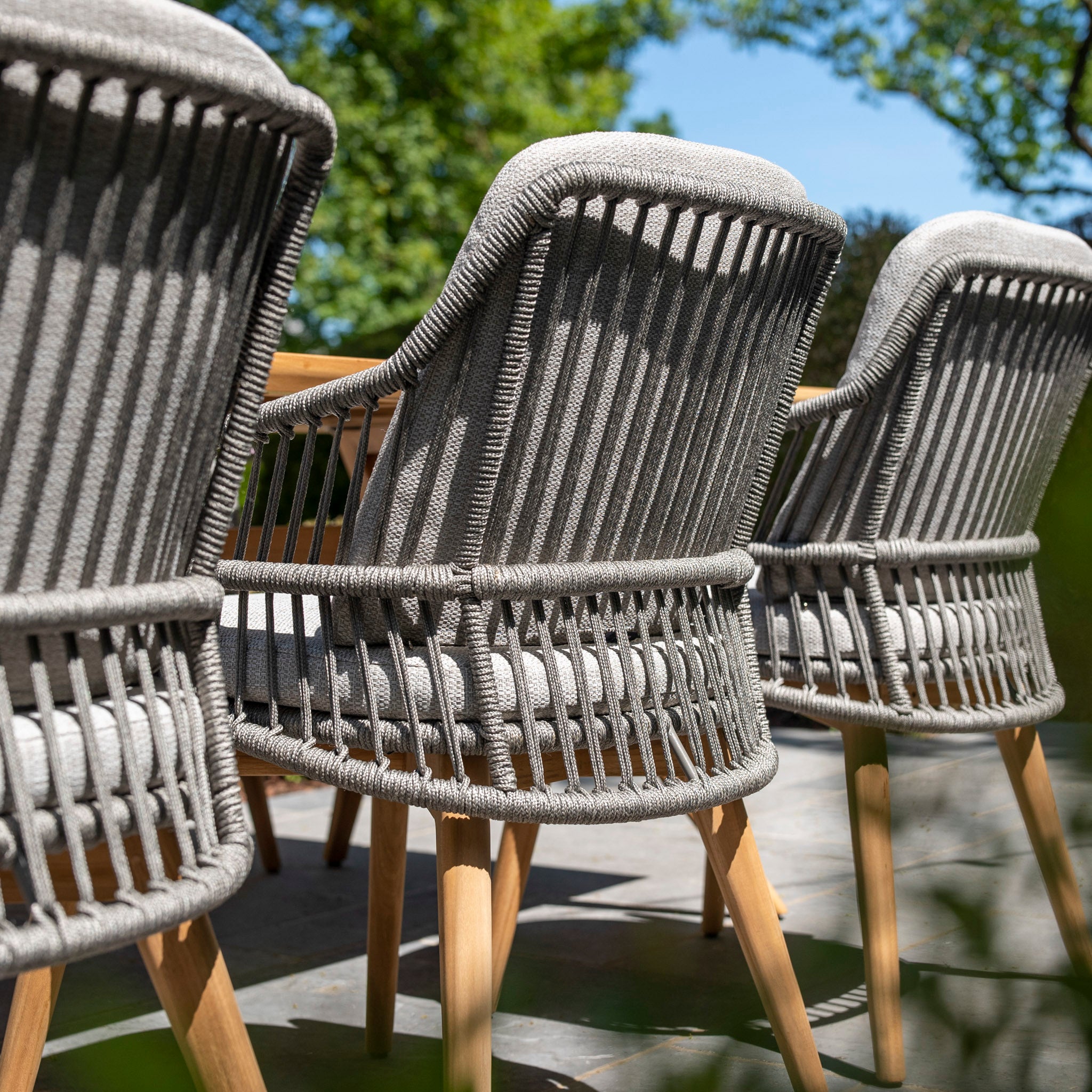 The Sempre 6 Seat Rectangular Dining Set with Teak Table in Light Grey is arranged on a sunny patio, featuring chairs with grey woven rope backs and teak wood legs.

