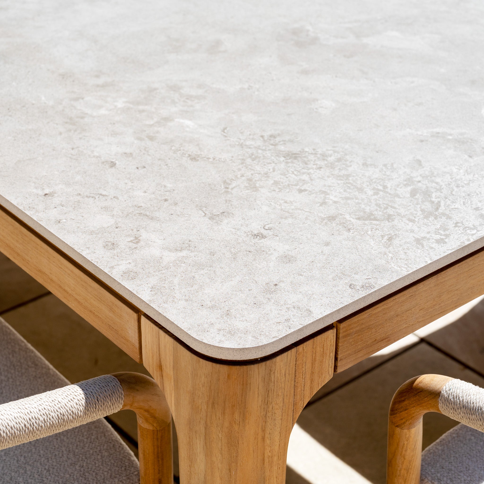 A close-up of the Siena 8 Seat Dining Set with Ceramic Top Table in Fawn, showing its light stone-finish surface with wooden edges and two teak wood chairs nearby.