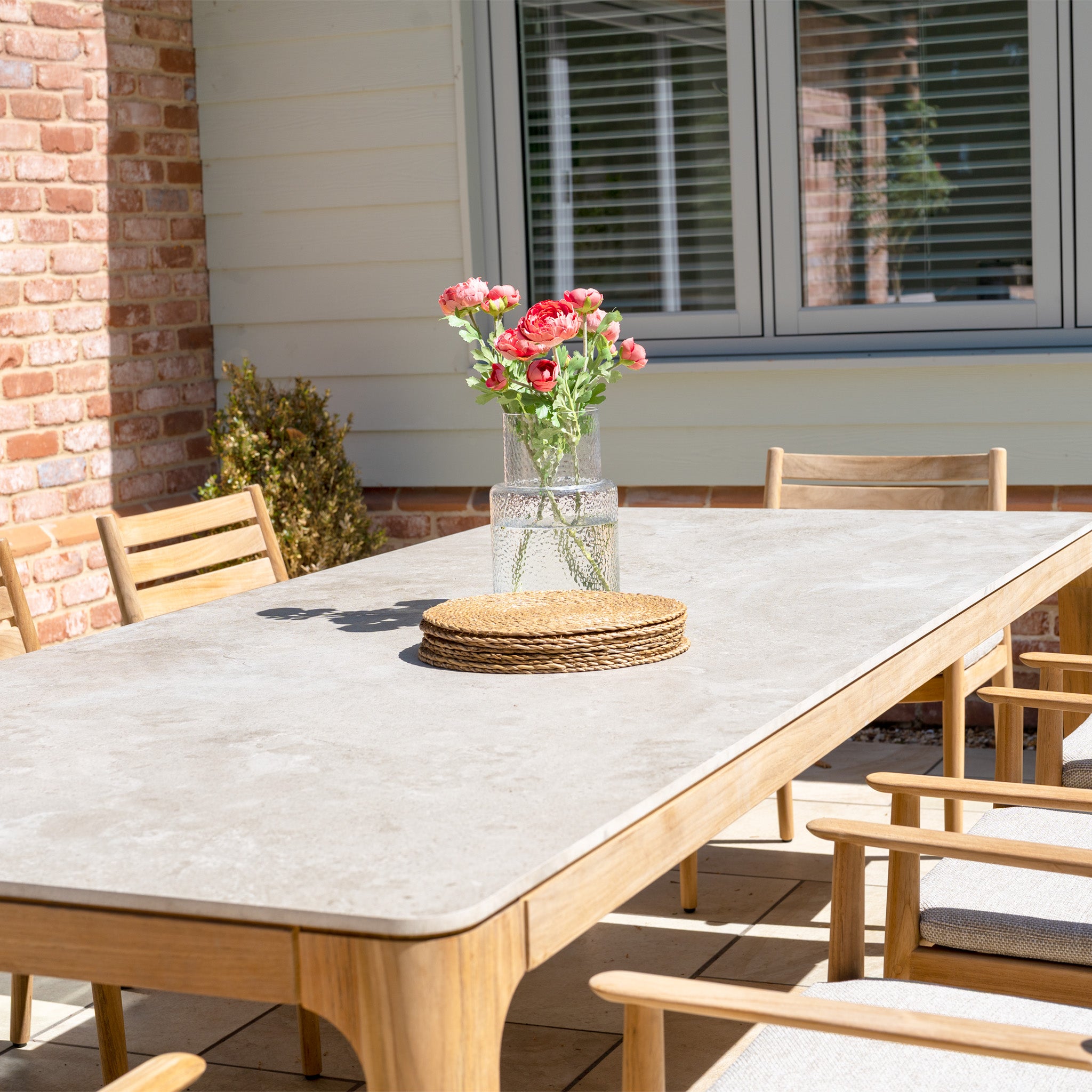 Siena 8 Seat Dining Set with Ceramic Top Table in Fawn—includes chairs and a spacious table, styled with a vase of pink flowers and round placemats, bathed in sunlight for an elegant dining experience.