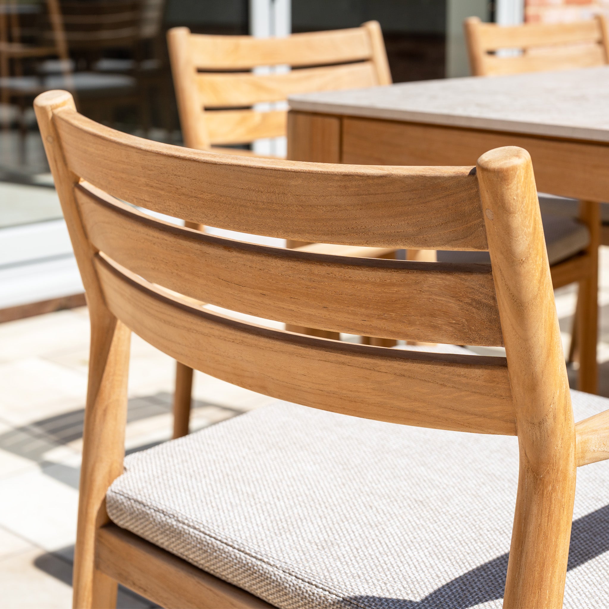Close-up of a teak wood outdoor chair with light cushion from the Siena 8 Seat Dining Set with Ceramic Top Table in Fawn, positioned next to the matching table and additional chairs.