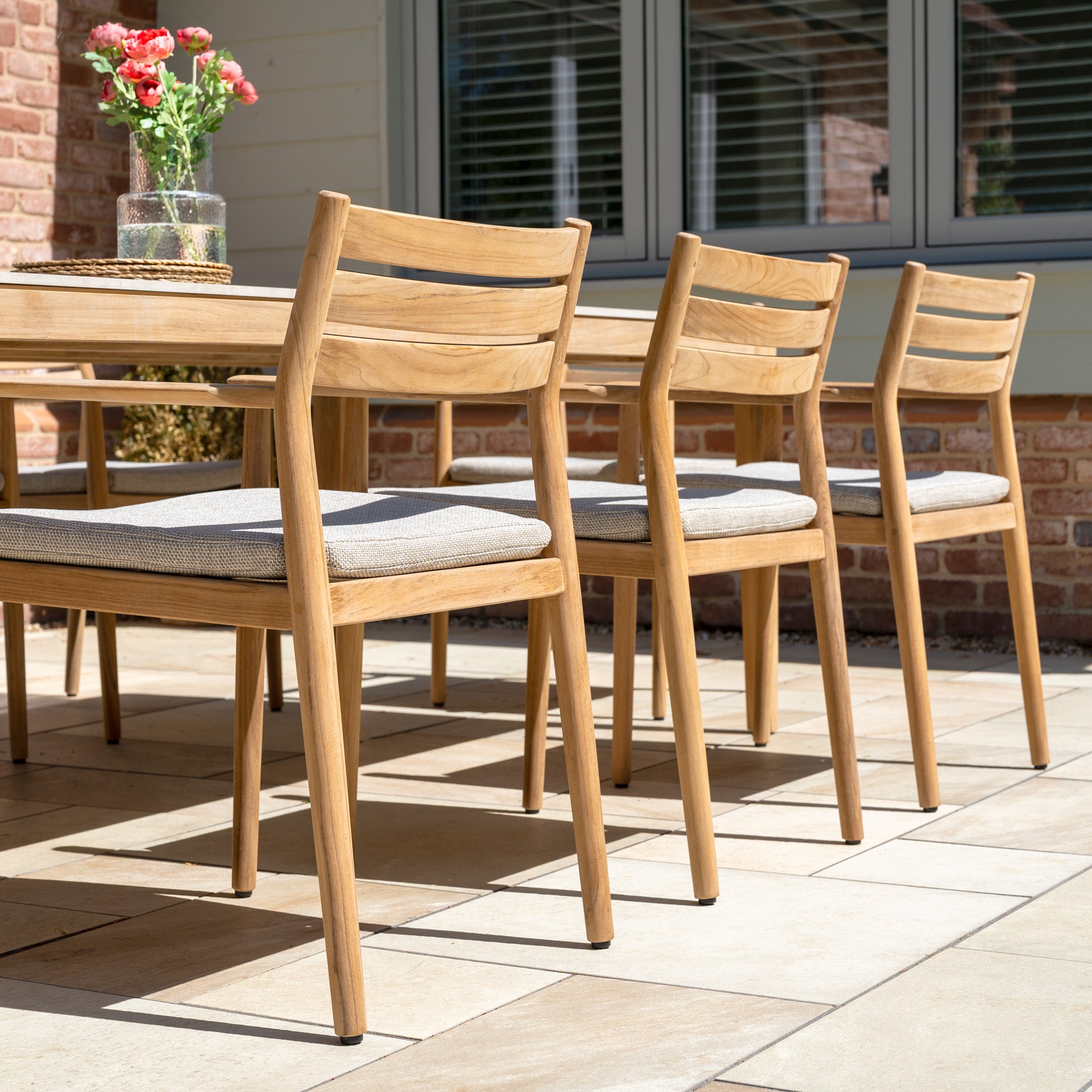 The Siena 8 Seat Dining Set with Ceramic Top Table in Fawn features cushioned teak wood chairs and a ceramic top table on a sunny patio; a vase of flowers adds charm to this inviting outdoor arrangement.
