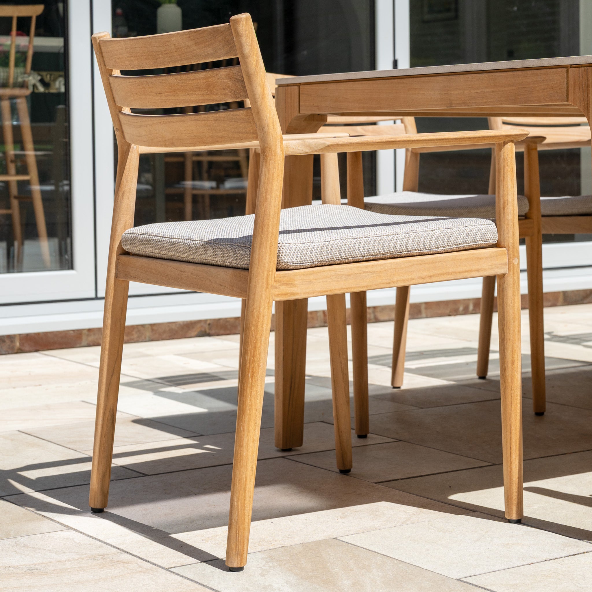 A light teak wood chair with a cushioned seat is placed next to the Siena 8 Seat Dining Set with Ceramic Top Table in Fawn, arranged on a tiled patio.