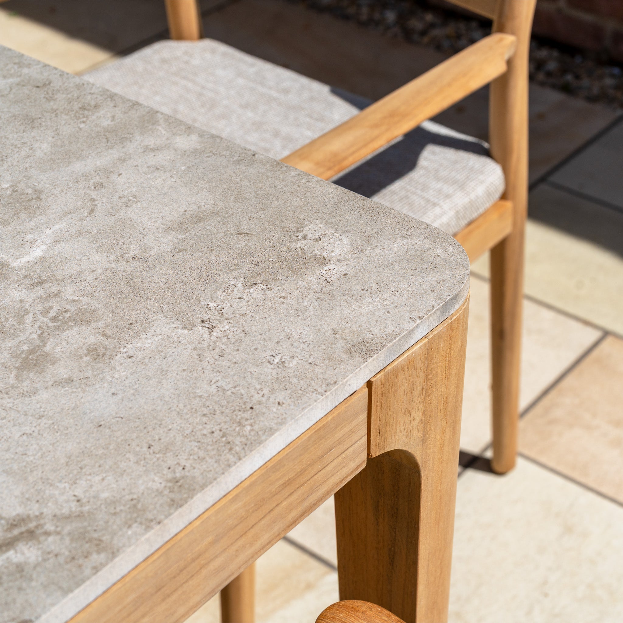 Close-up of the Siena 8 Seat Dining Set with a ceramic top table in fawn and teak wood legs, beside a cushioned wooden chair on a tiled patio.