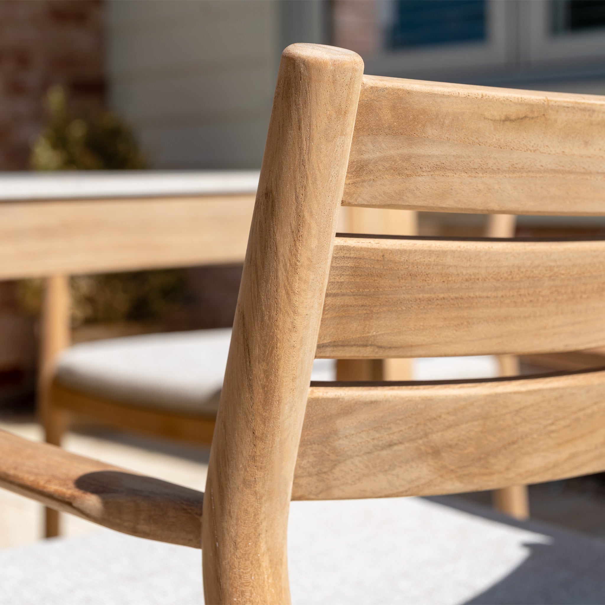 Close-up of a teak wood chair with a curved backrest, outdoors in sunlight, alongside the Siena 8 Seat Dining Set with Ceramic Top Table in Fawn.