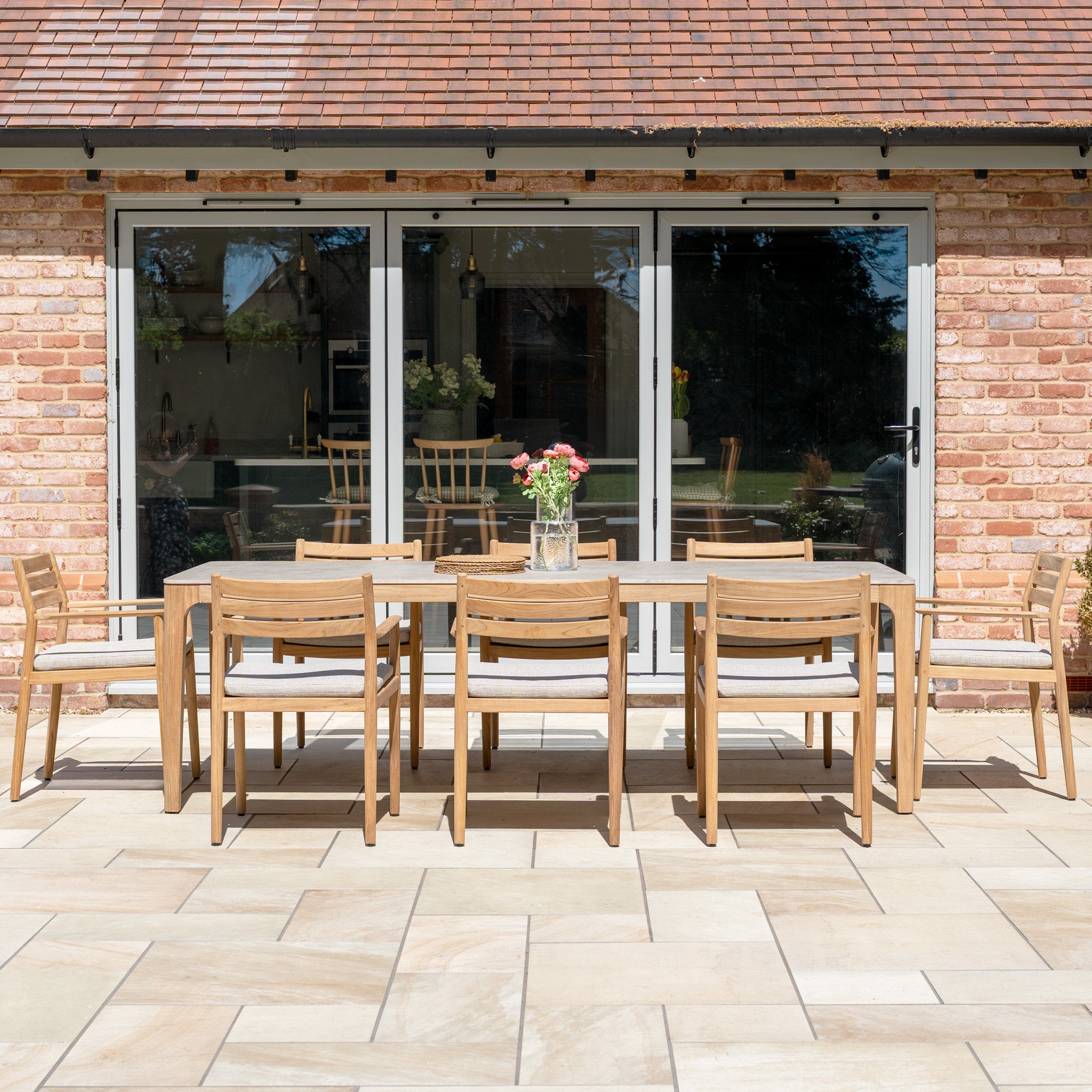 The Siena 8 Seat Dining Set with Ceramic Top Table in Fawn is arranged on the patio in front of glass doors, adorned with a vase of flowers.