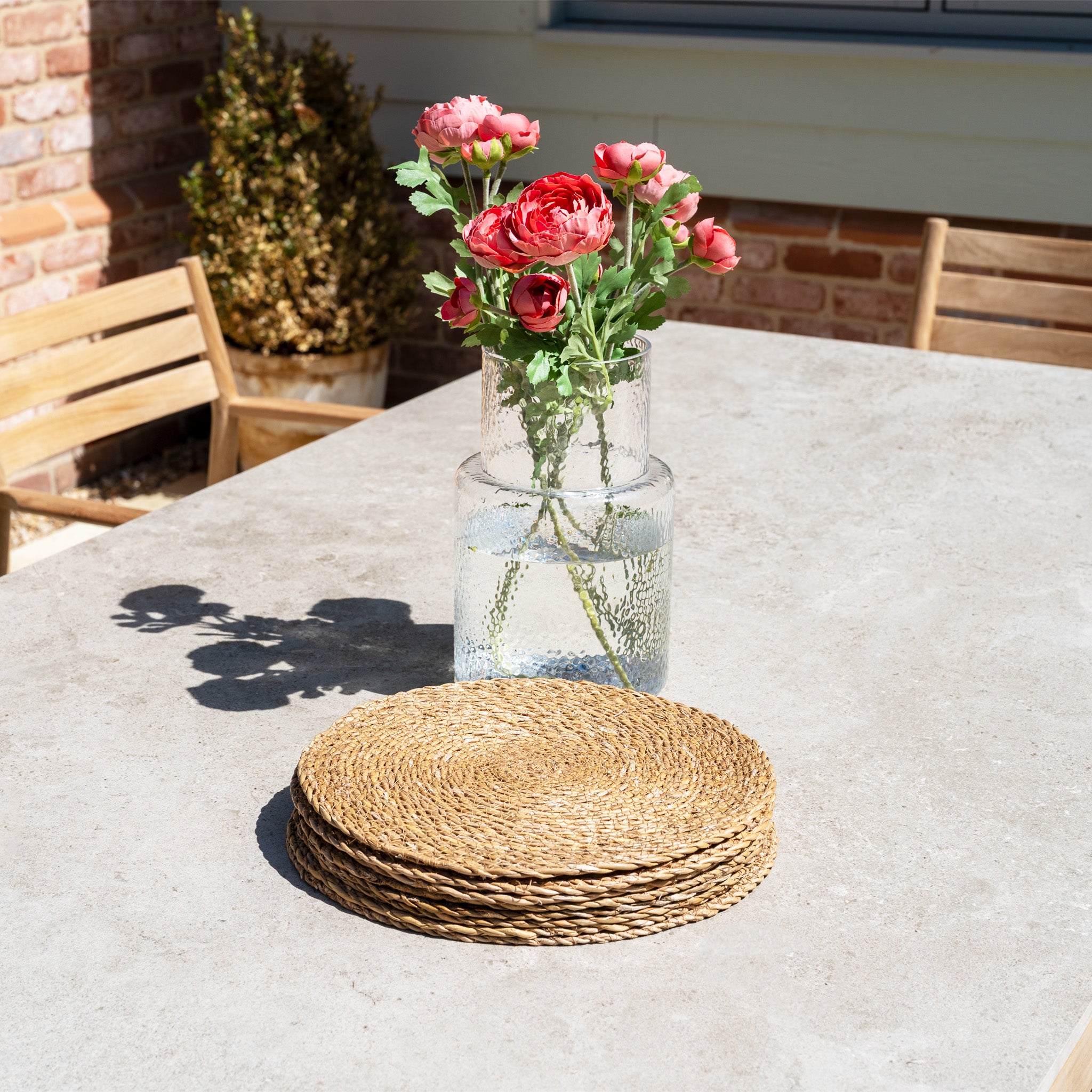 A vase with pink flowers and wicker placemats rests on the Siena 8 Seat Dining Set with Ceramic Top Table in Fawn, illuminated by sunlight outdoors.