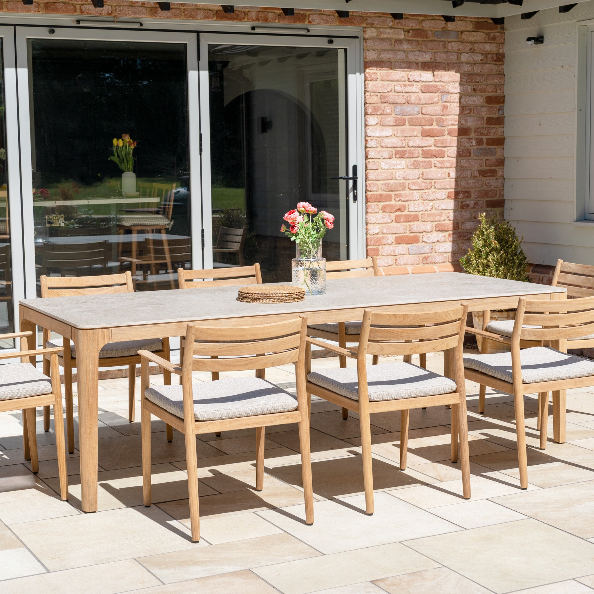 The Siena 8 Seat Dining Set with Ceramic Top Table in Fawn, arranged on a tiled terrace, features eight chairs and a stylish table adorned with a vase of flowers.