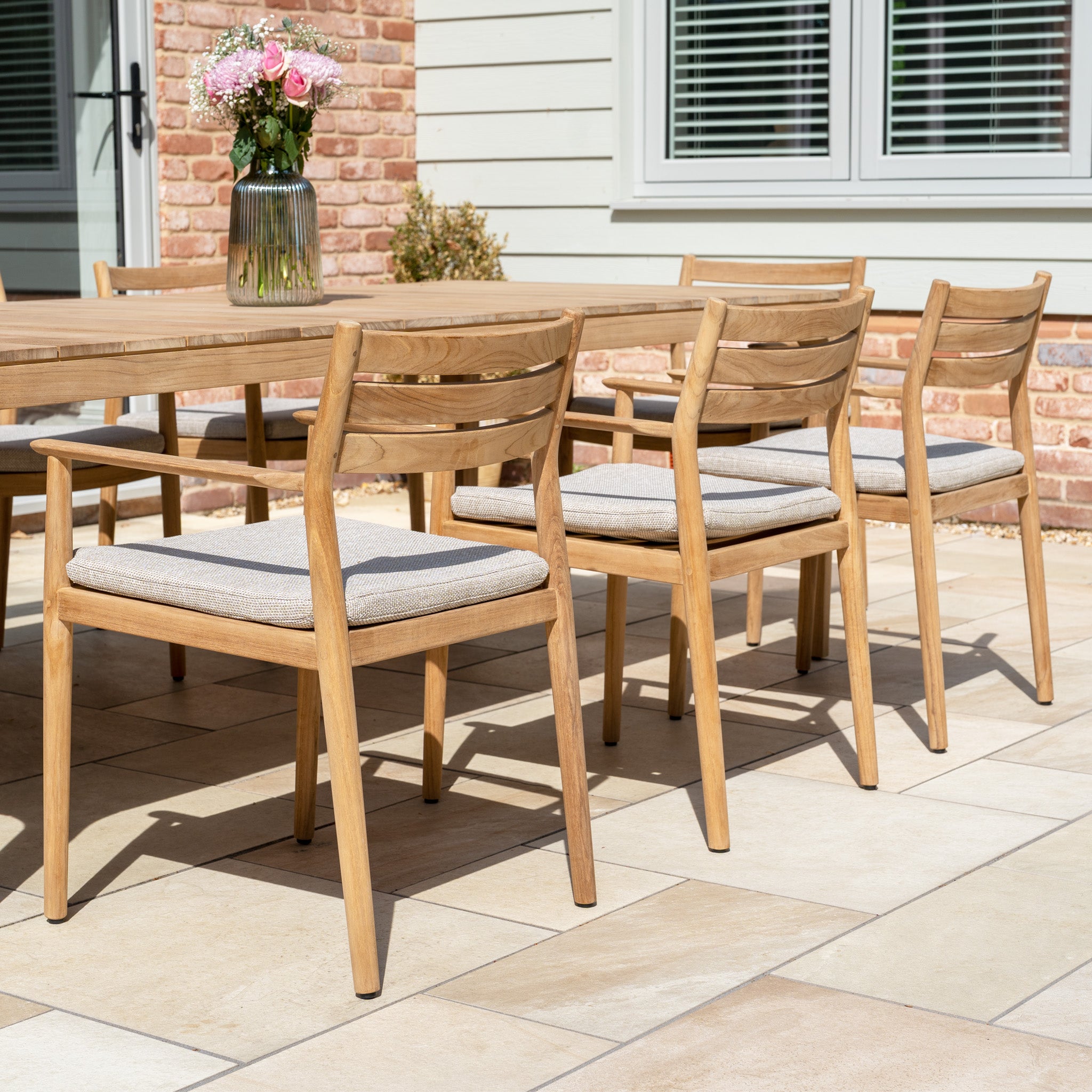 The Siena 8 Seat Dining Set with Teak Table in Fawn, featuring matching chairs, is arranged on a tiled patio near the house—ideal outdoor furniture for relaxing or entertaining with family and friends.