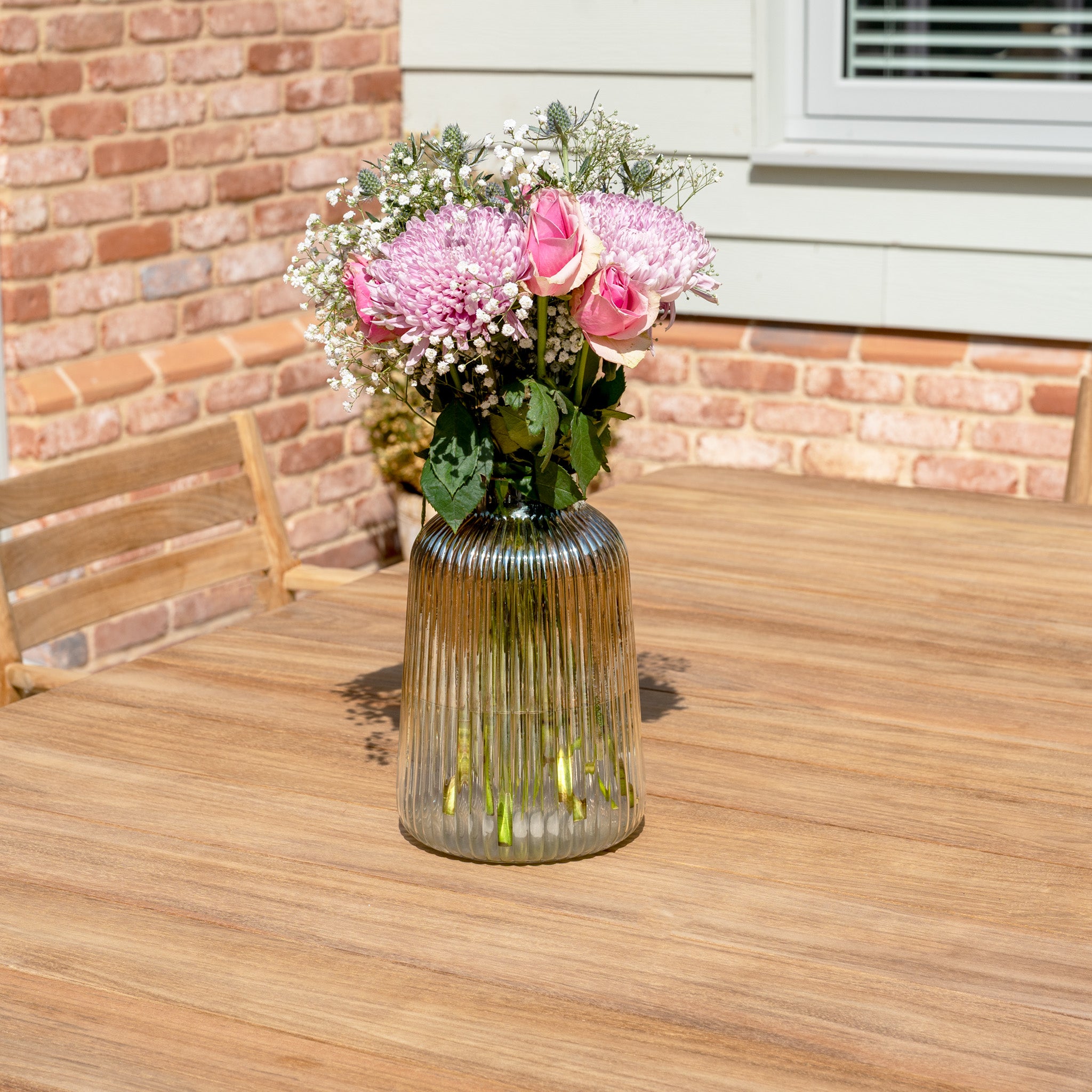 A glass vase with pink flowers rests on the Siena 8 Seat Dining Set with Teak Table in Fawn, surrounded by outdoor chairs.