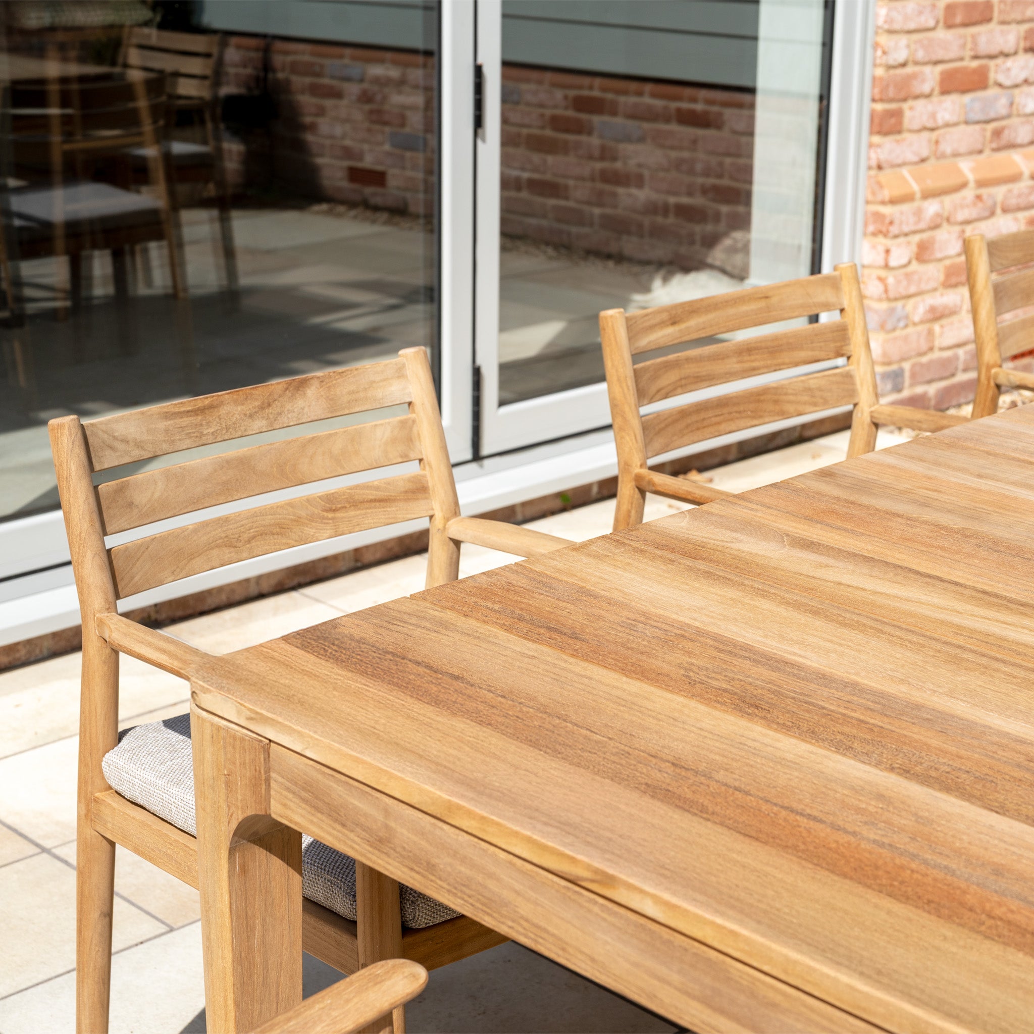 The Siena 8 Seat Dining Set with Teak Table in Fawn creates an inviting outdoor arrangement near glass doors and a brick wall, bathed in sunlight.